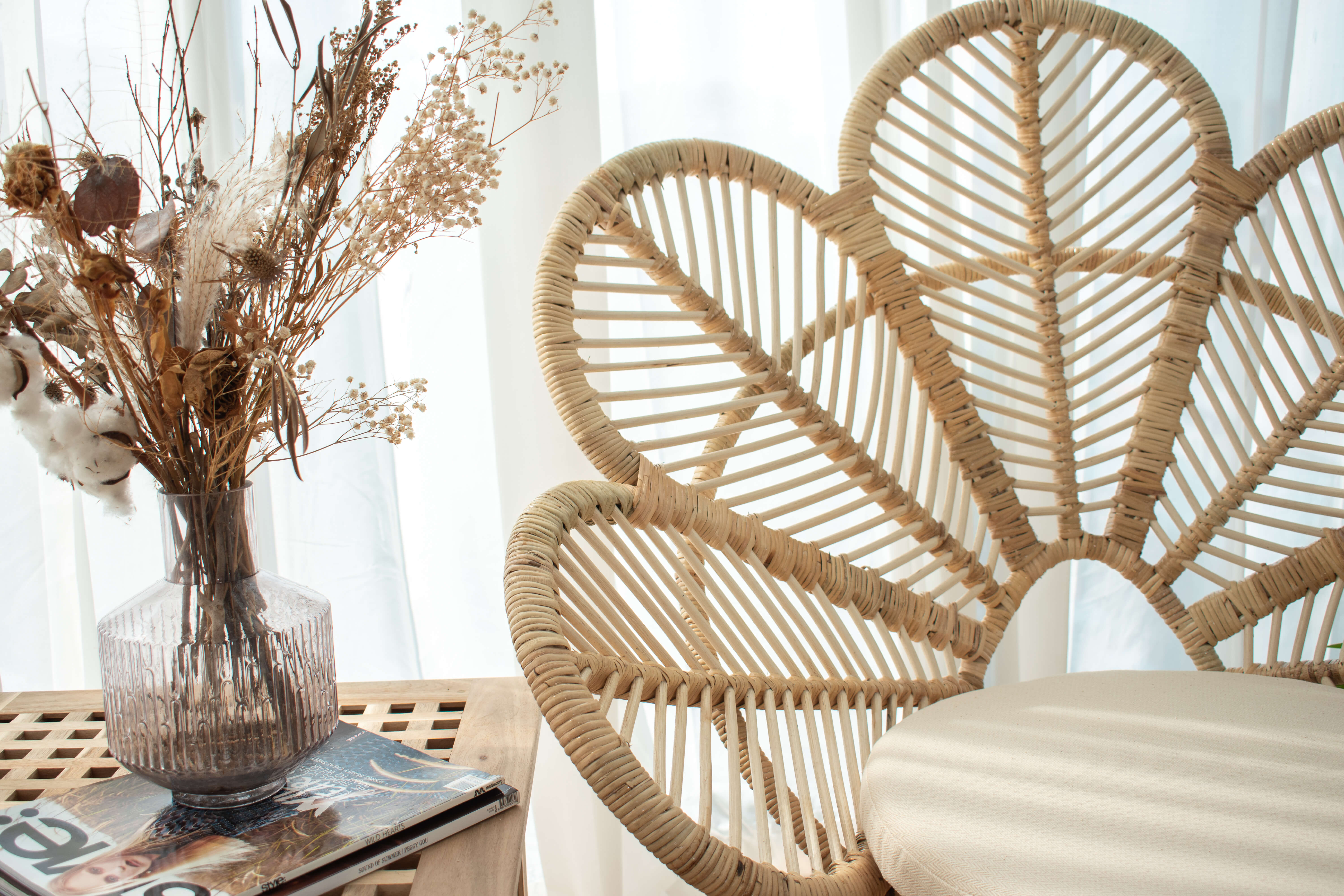 A Natural White Rattan Leaf Chair with a fan-shaped backrest next to a wooden table holding a glass vase filled with dried flowers and a magazine.