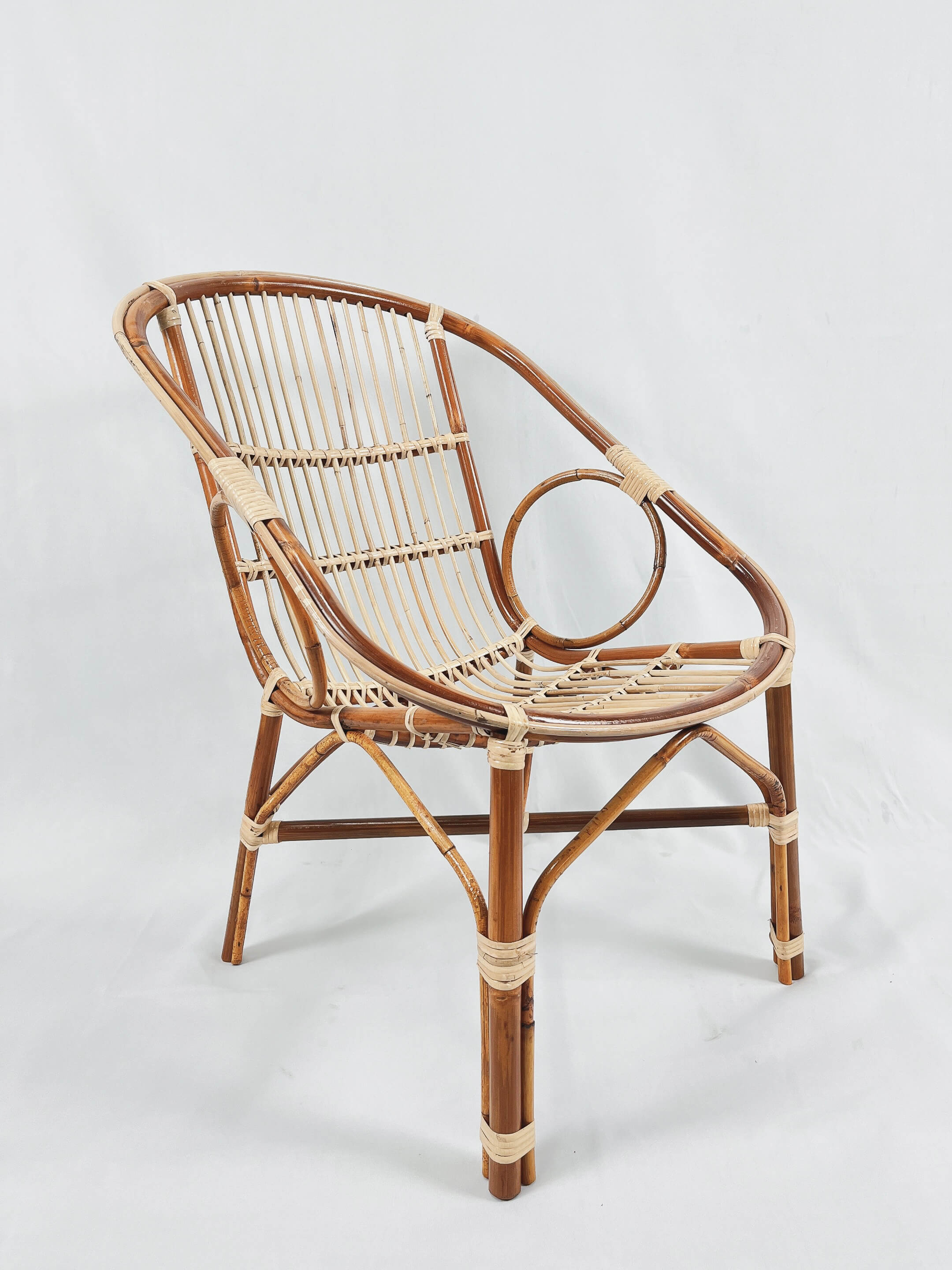 The Two Ring Chair, crafted from brown wicker with curved armrests and featuring a circular pattern in the backrest, is set against a plain white background.