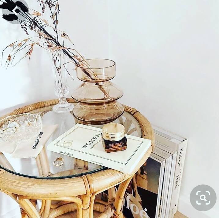 A RC Rattan Table With Glass Top holds a glass vase with dried flowers, a glass dish, a book titled "WOMEN", and a jar of cream. Additional books are stacked beside the table.