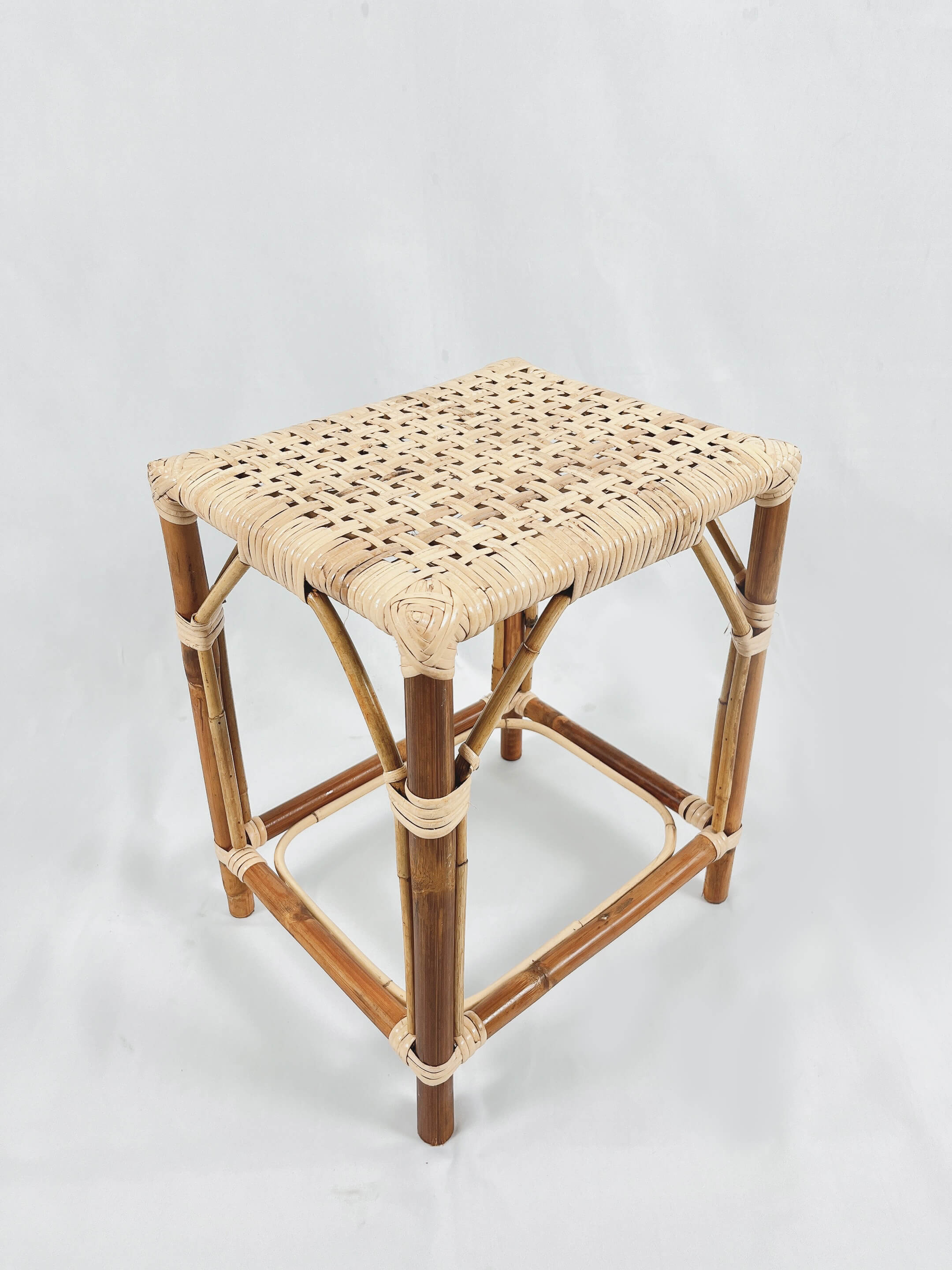 The RC Sewing Stool is a compact square stool crafted from bamboo, featuring a woven wicker seat, styled against a minimalist white backdrop.