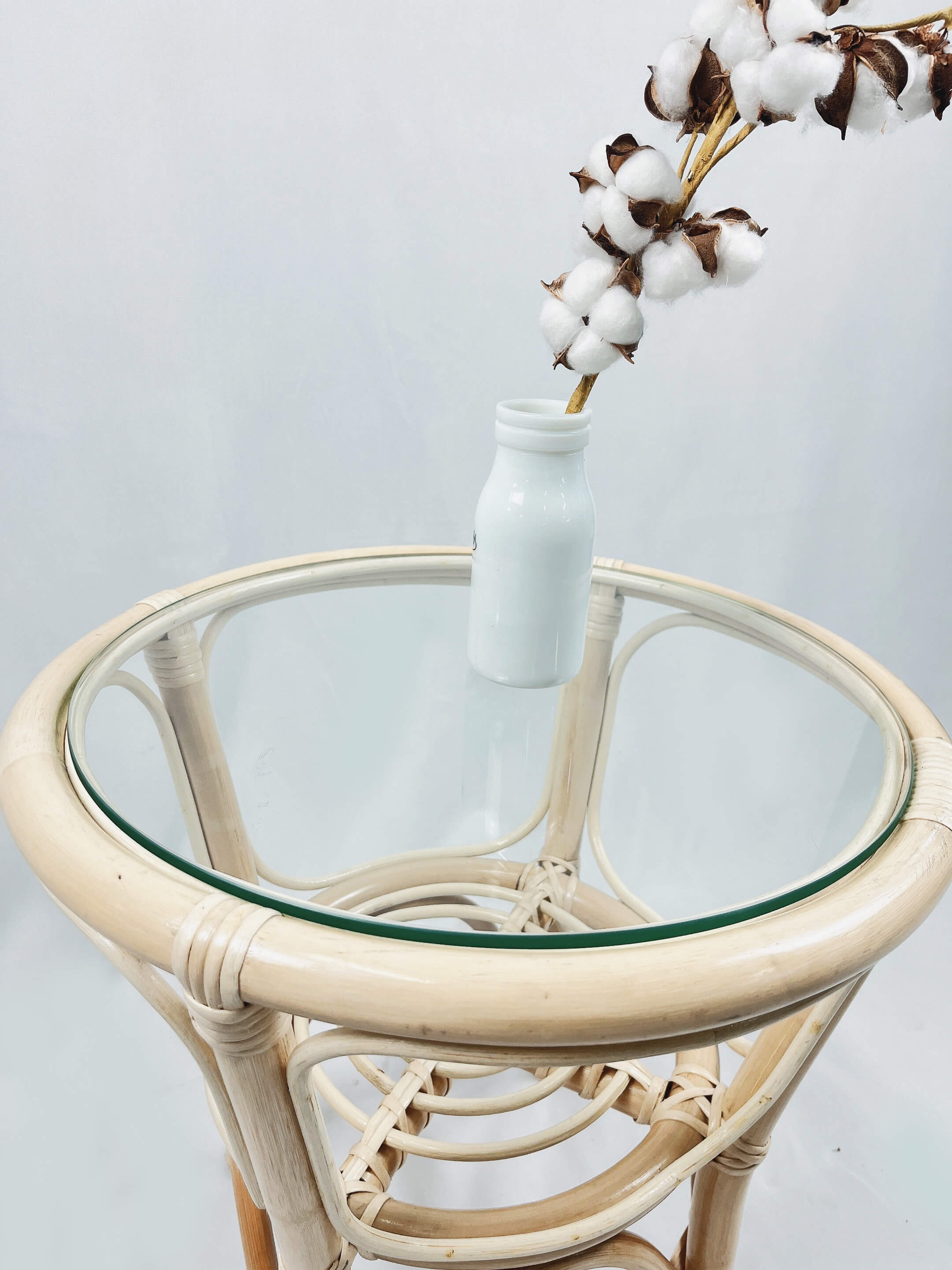 A round RC Rattan Table with a glass top, complemented by a white vase holding a branch of cotton flowers.