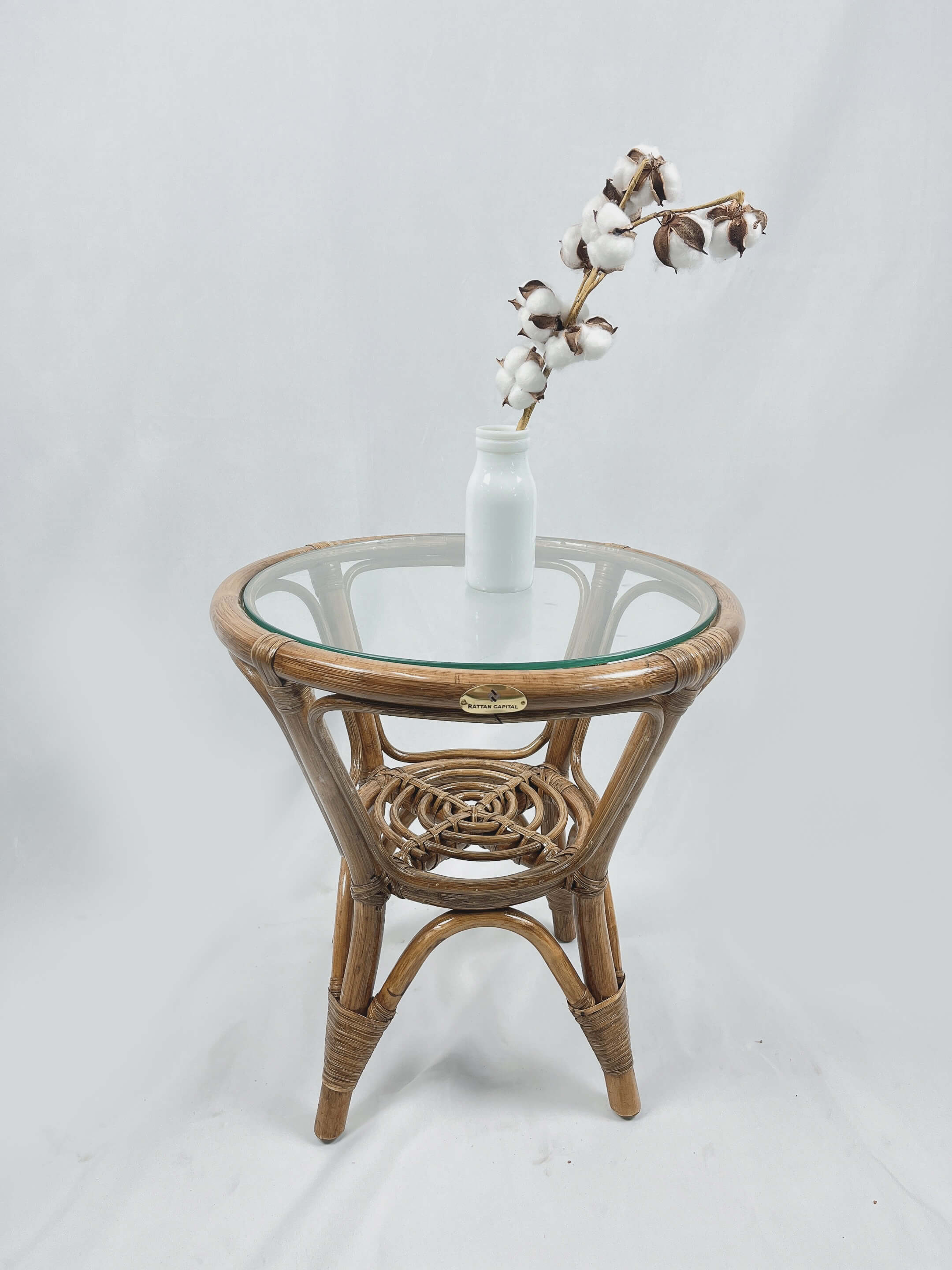 An RC Rattan Table With Glass Top holds a white vase containing white cotton stems.