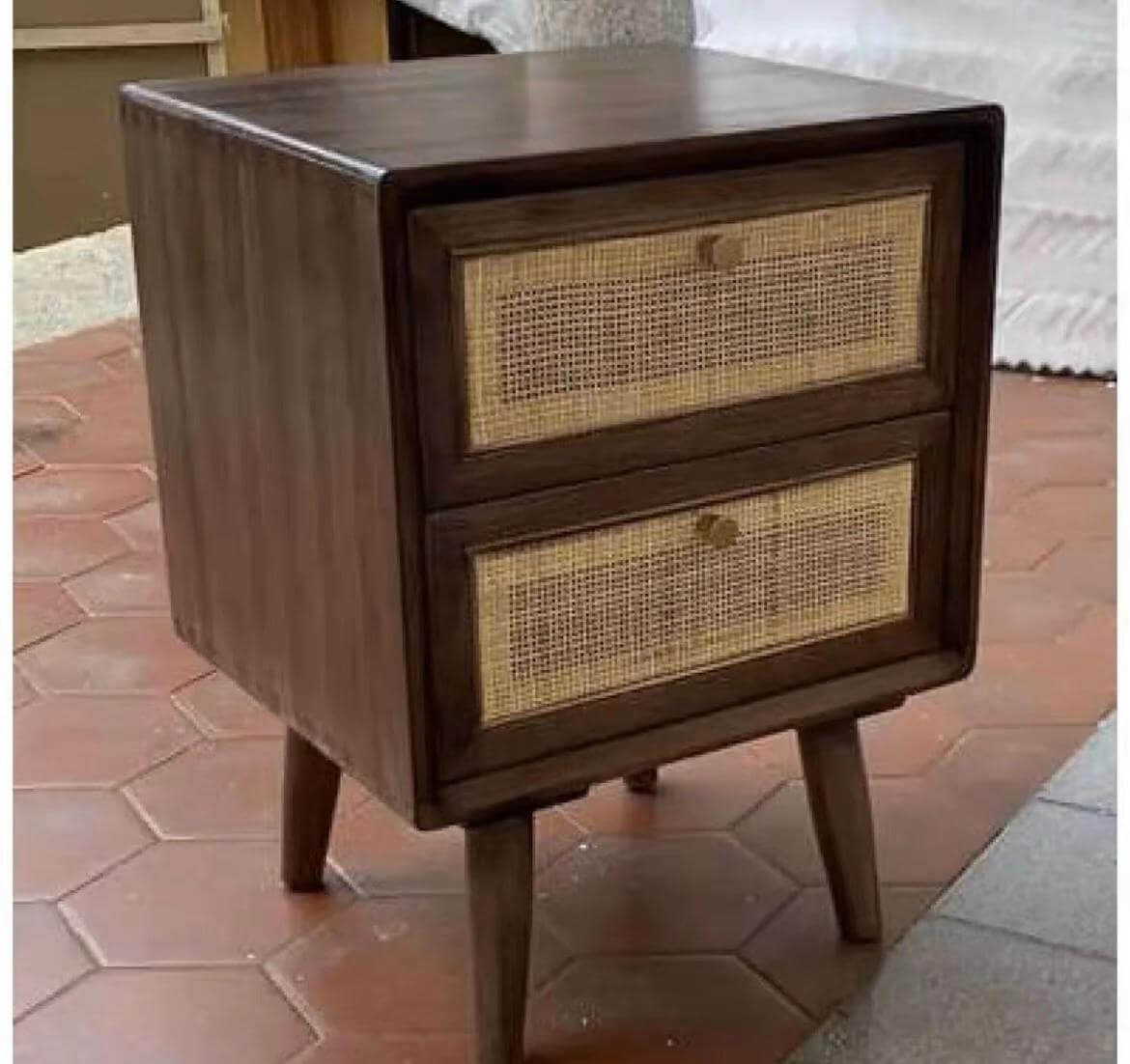 The Raffles Side Table with two drawers featuring cane webbing fronts stands on four angled legs on a tiled floor.