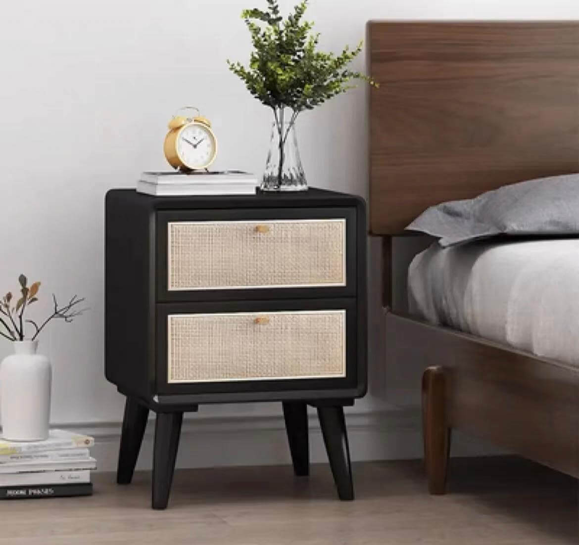 The Raffles Side Table, a black wooden nightstand with two woven drawers, sits next to a wooden bed. It has a plant, an alarm clock, and a book on top. A white vase with twigs is on the floor beside it.