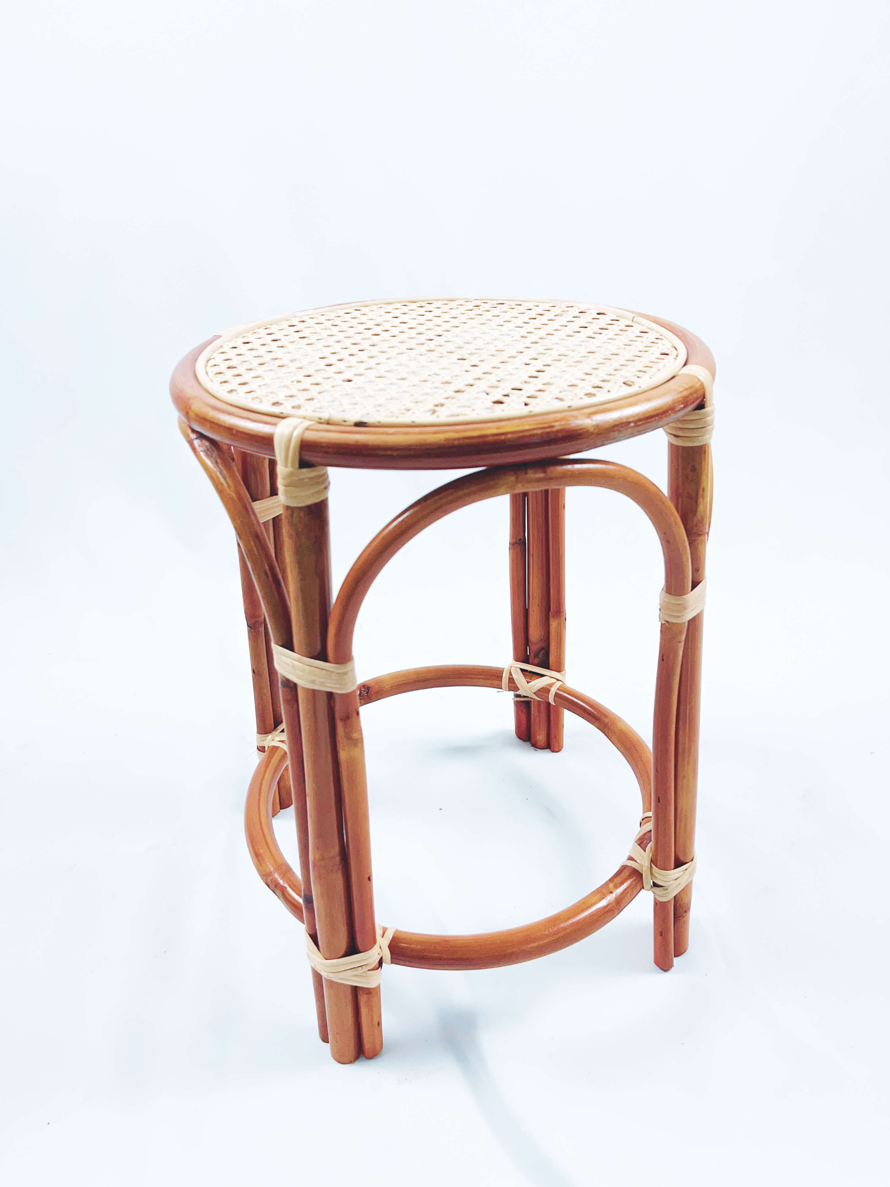 The RC Bar Stool (No Backframe) is a small, round stool with a woven rattan seat and wooden legs, featuring supporting circular braces.