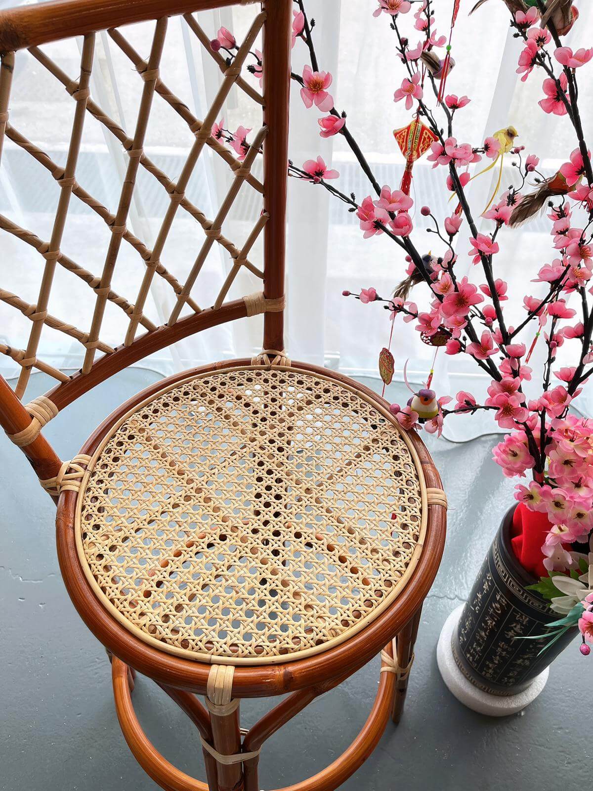 The PO - RC Bar Stool (With Backframe) is positioned next to a vase holding pink cherry blossoms and adorned with a butterfly decoration. The background is complemented by a light-colored curtain.