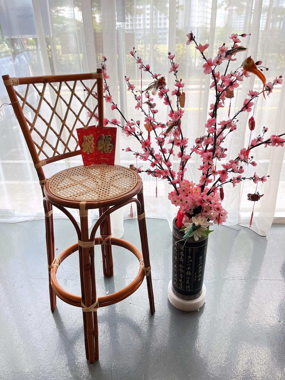 A PO - RC Bar Stool (With Backframe) is placed beside a potted artificial cherry blossom tree on a floor by a window. Two red envelopes with gold Chinese characters rest on the stool.
