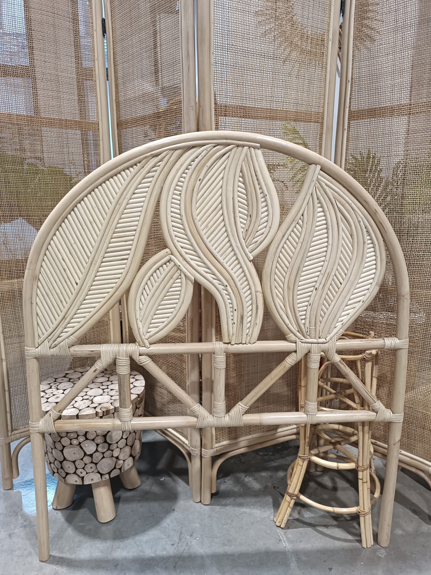 The Artistic Natural Leaf Rattan Single Headboard (Clearance) with leaf patterns is displayed in front of a wicker room divider, beside a small log stool and a bamboo stool.
