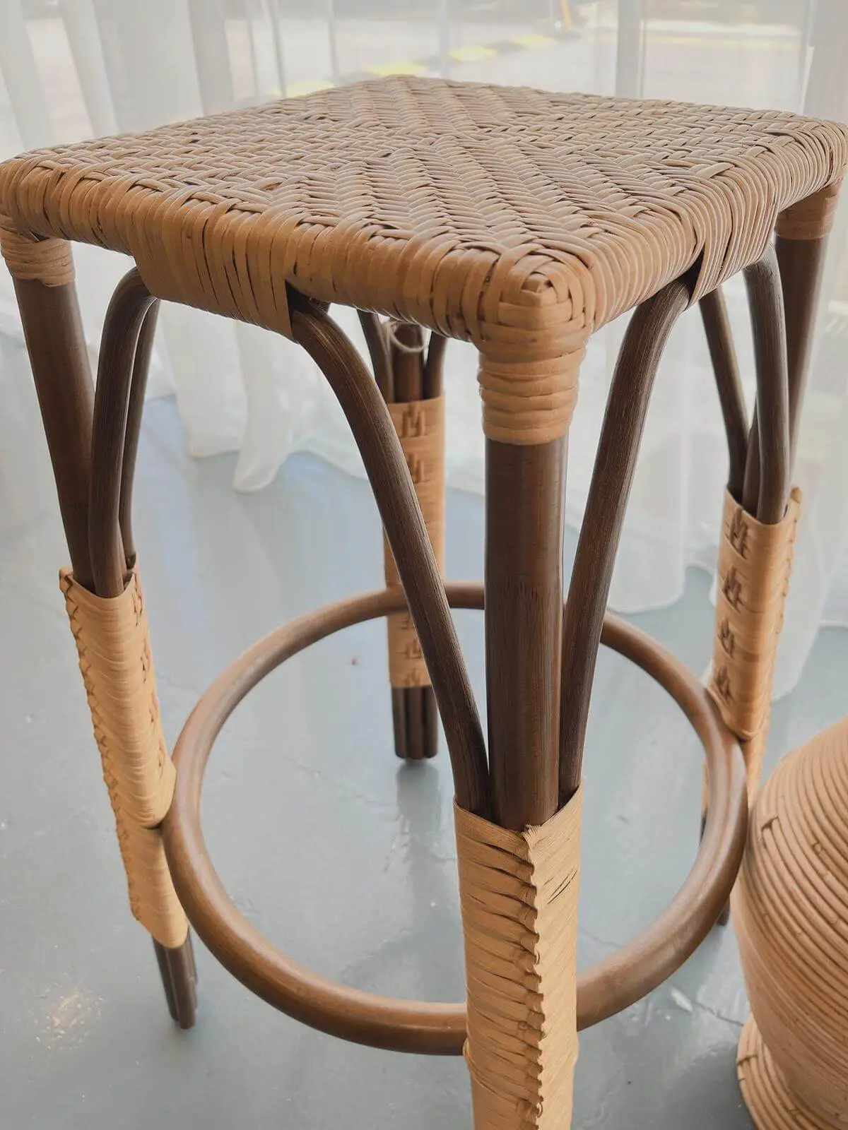 A square wicker stool with a woven seat and wooden legs, featuring circular support and woven accents. The stool is positioned on a pale blue floor.