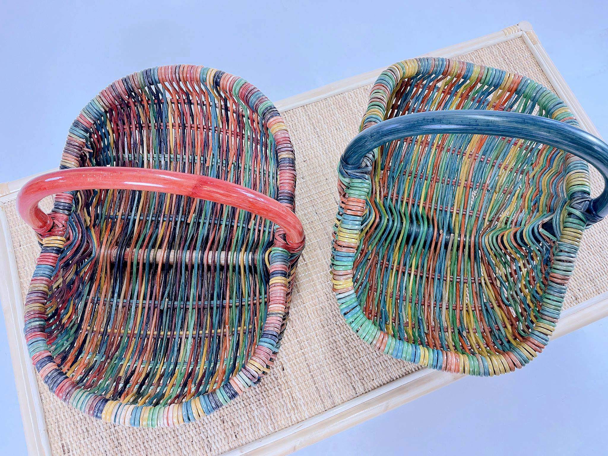 Two vibrant Wicker Basket LK10s, each featuring curved handles—one adorned with a red handle and the other with a blue handle—are placed side by side on a woven mat.