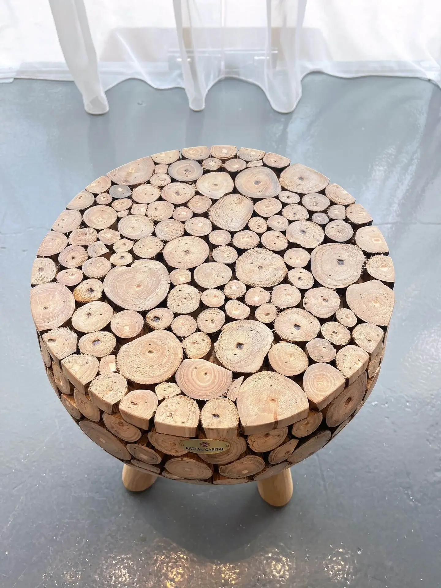 The Mosaic Teak Art Stool, featuring a top surface crafted from various sized circular teak cuts, stands on a grey, glossy floor near a white curtain.