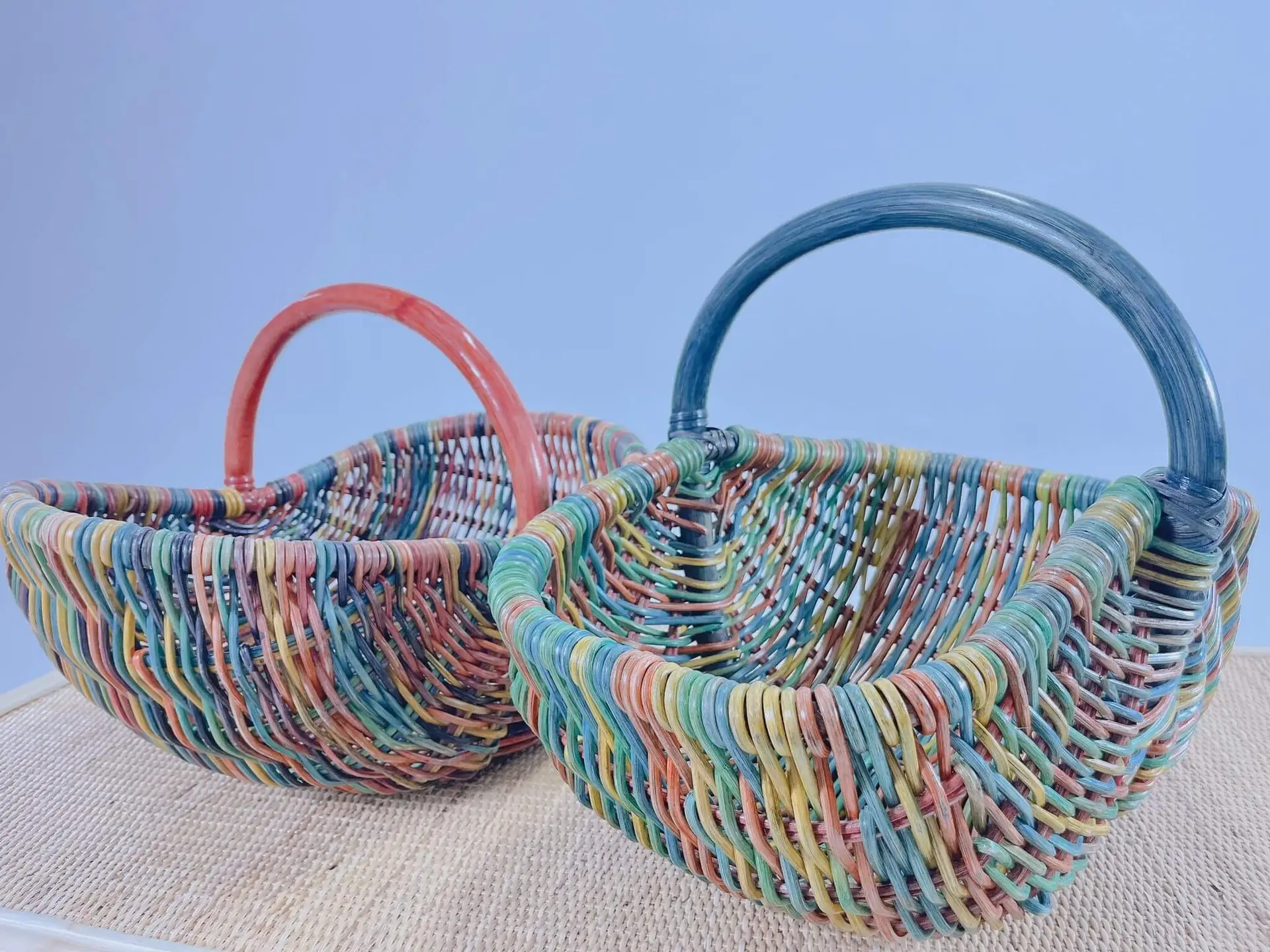 Two of Dawn's Plant Pots, multicolored and woven with curved handles—one featuring a red handle and the other a blue handle—placed on a beige surface.