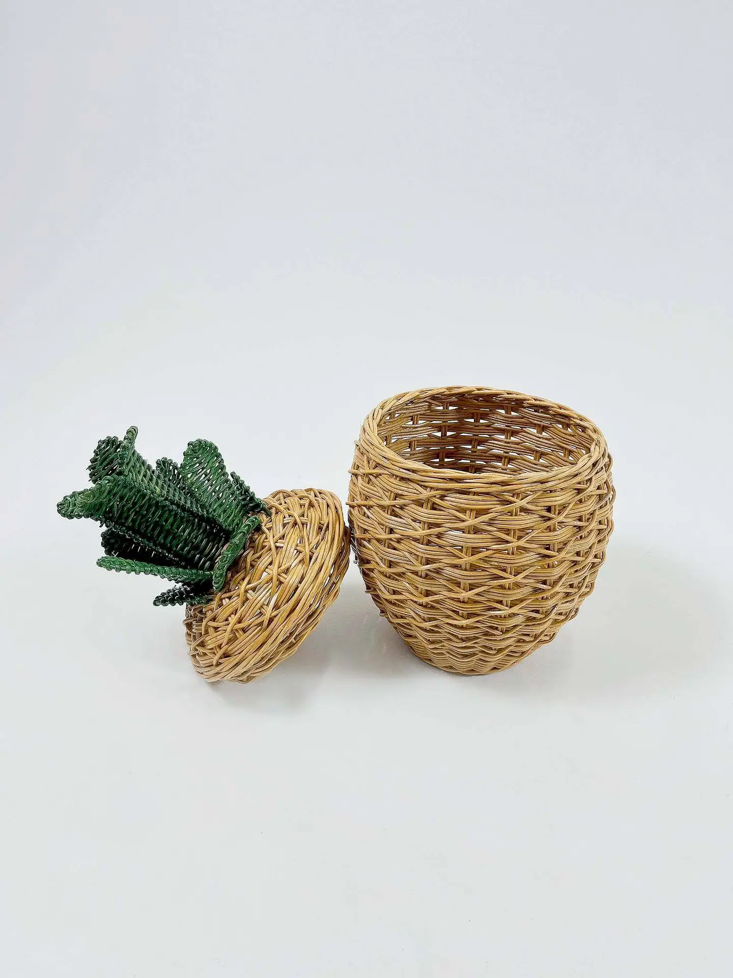 A small woven basket shaped like a pineapple, with the lid that includes green leaves resting beside it.