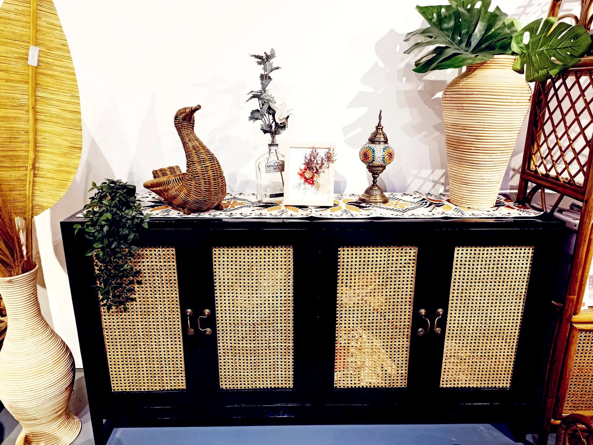 A black and wicker console table displays decor items including a wicker duck, a small tree, a framed picture, a lantern, and a large vase with green leaves on the right side.