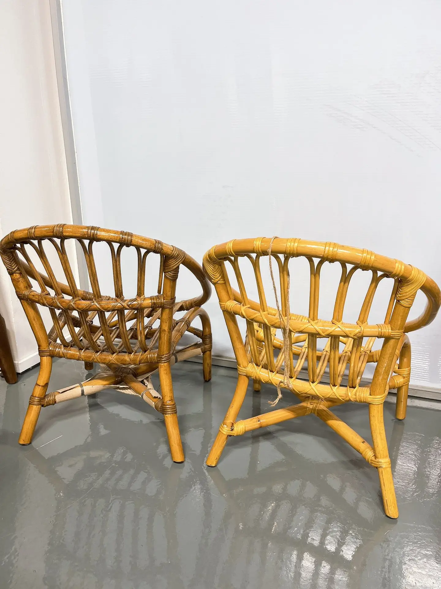Two wicker chairs with curved backs and wooden frames are placed on a shiny gray floor, positioned facing a white wall.