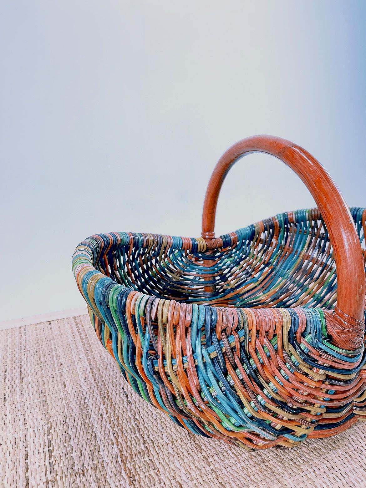 The Wicker Basket LK10, featuring a woven multicolored design and a convenient handle, rests on a textured beige surface against a light background.