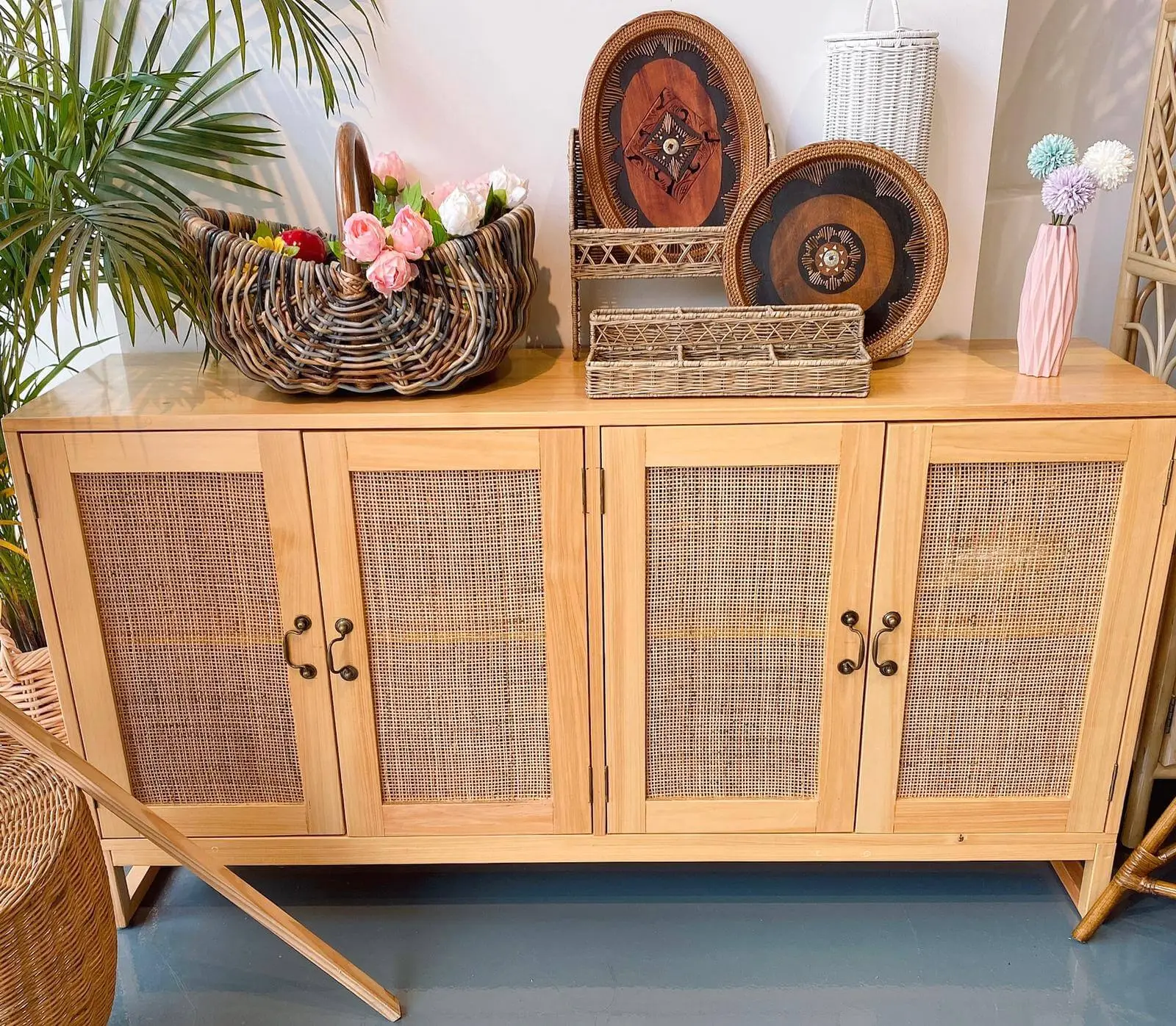 A wooden cabinet with wicker doors and decorative handles is topped with various woven baskets, plates, and faux flowers, positioned against a wall with plants on both sides.