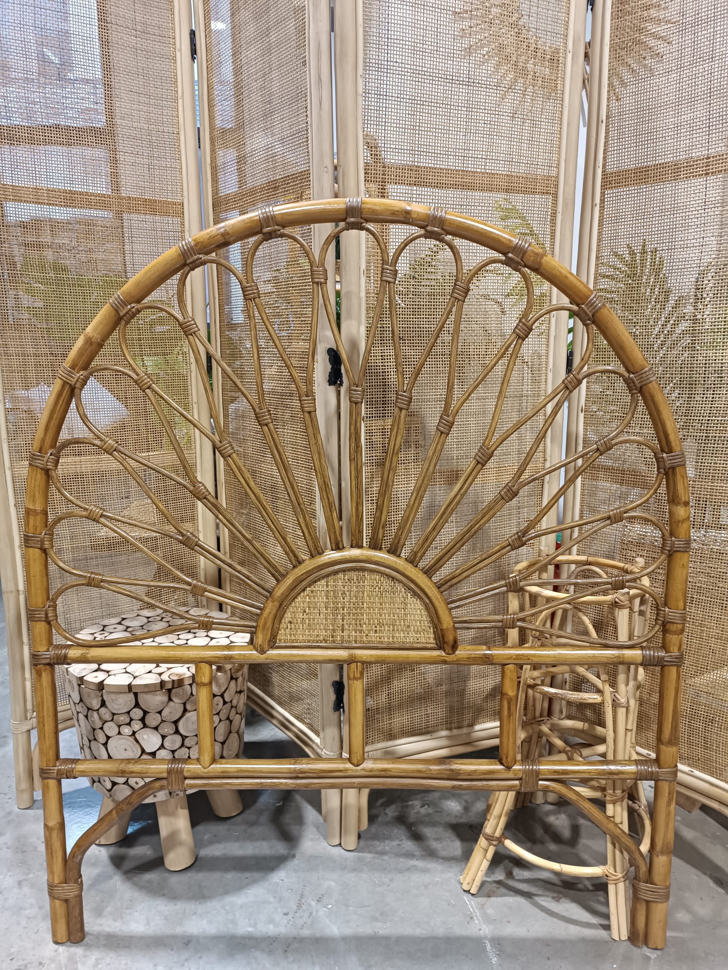 The Floral Natural Rattan Headboard (Clearance) is shown in front of a woven room divider, alongside a round log-patterned stool and a matching rattan side table. The Floral Natural Rattan Headboard (Clearance) is shown in front of a woven room divider, alongside a round log-patterned stool and a matching rattan side table.