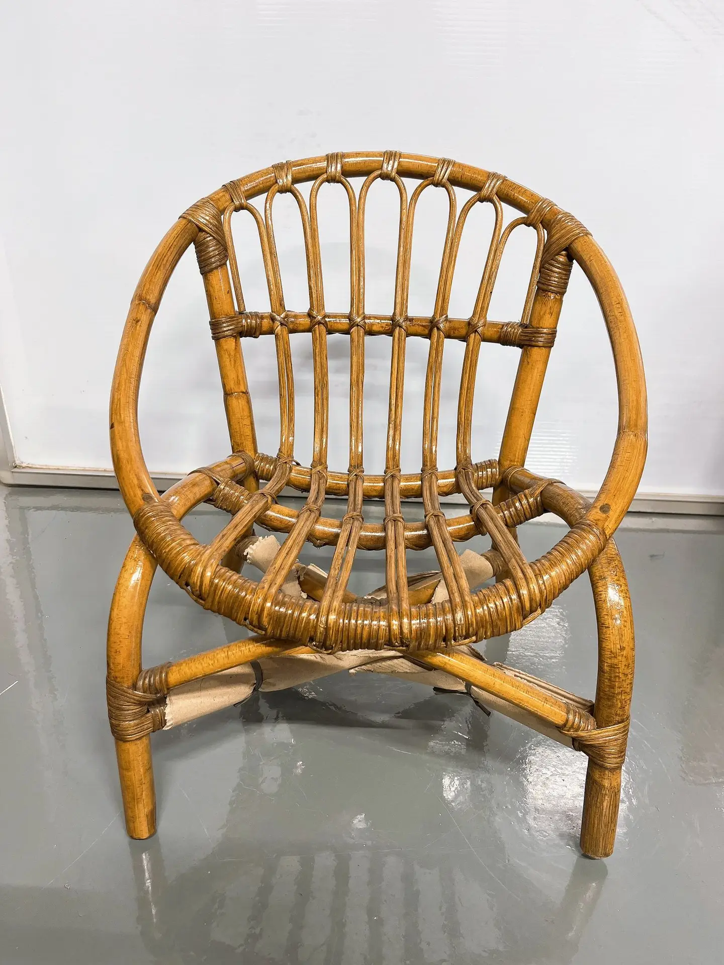 A small, round, wicker chair with a wooden frame and open back design, placed on a gray reflective floor against a white background.