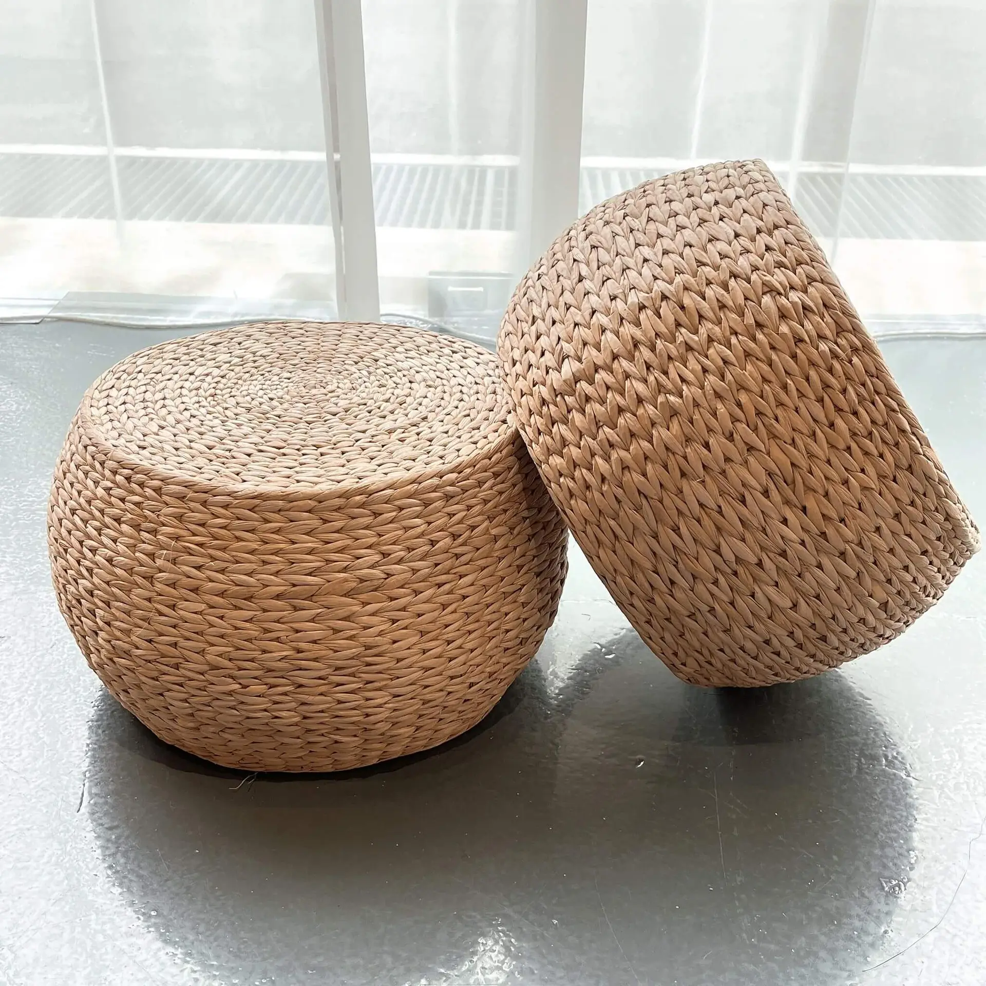 Two round Seagrass Ottomans, one resting upright and the other tilted, on a smooth, grey floor next to windows.