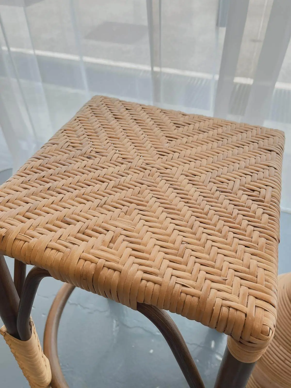 A close-up view of the woven top of a wooden stool with a checkered pattern. The stool is placed near a window with sheer, white curtains.