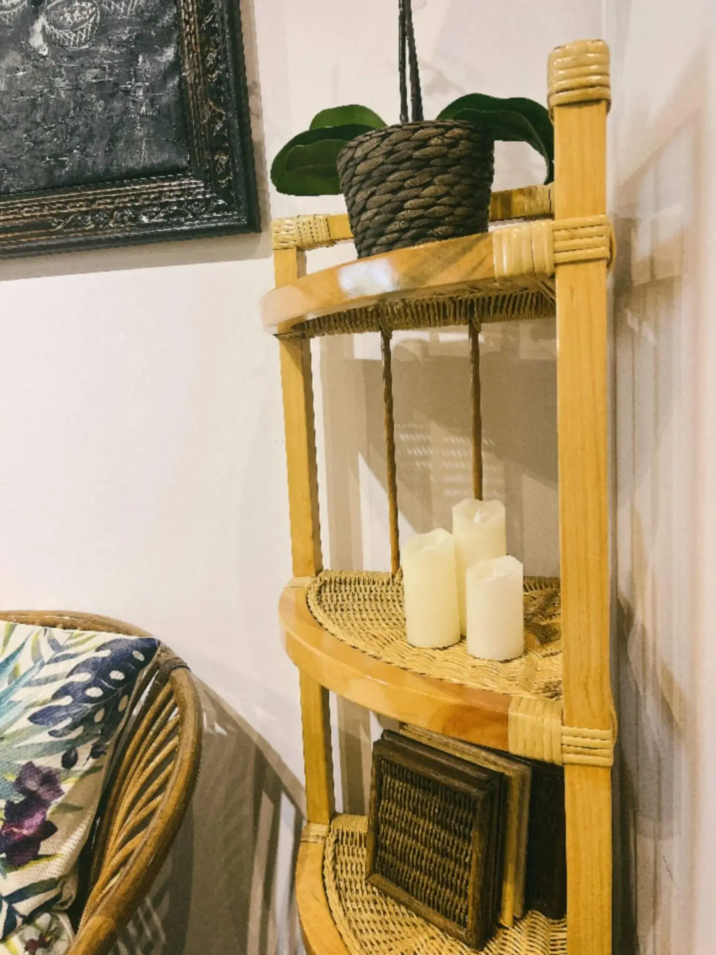 A small wooden corner shelf set holds three white candles, a potted plant, and some books, next to a rattan chair with a floral patterned cushion.