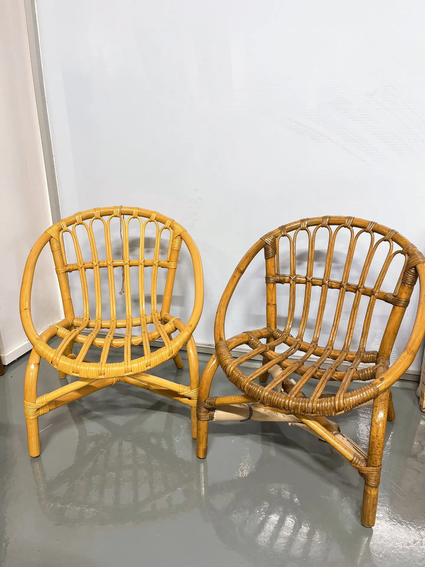Two rattan chairs with curved backs and open, woven designs are placed side by side on a smooth, gray floor against a white wall.