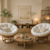 Two Manau Papasan chairs with natural frames and cushions, plus a small wooden table with vases and cups, sit in a minimalist, neutral-toned living room decorated with plants and simple accents.