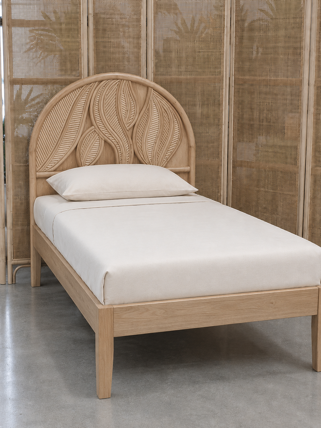 A single bed featuring the Artistic Natural Leaf Rattan Single Headboard (Clearance) with beige bedding stands on a concrete floor, set before a woven rattan room divider.