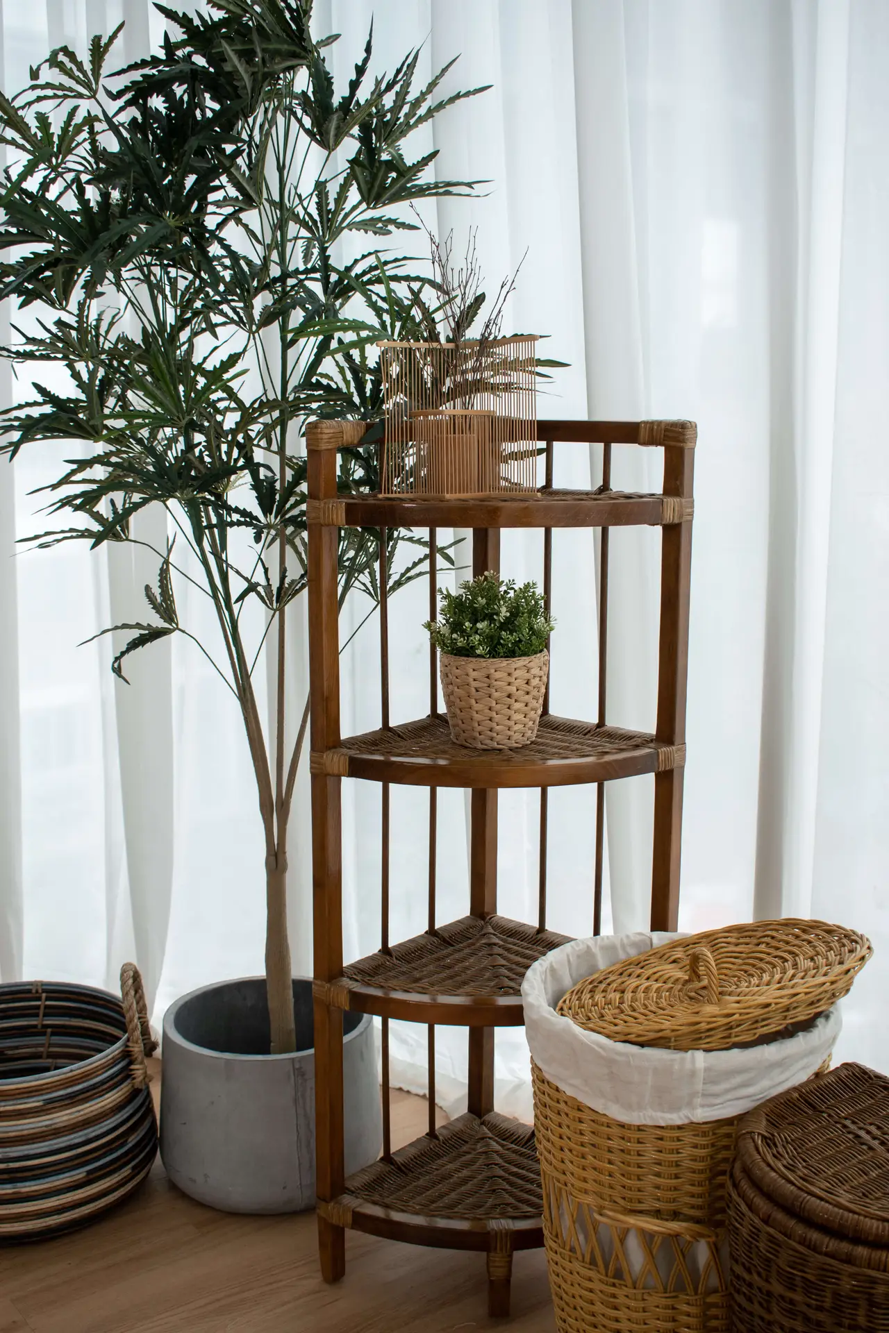 A Wicker Rack stands against white curtains, holding a potted plant and decor items. Nearby are baskets and a tall plant in a pot.