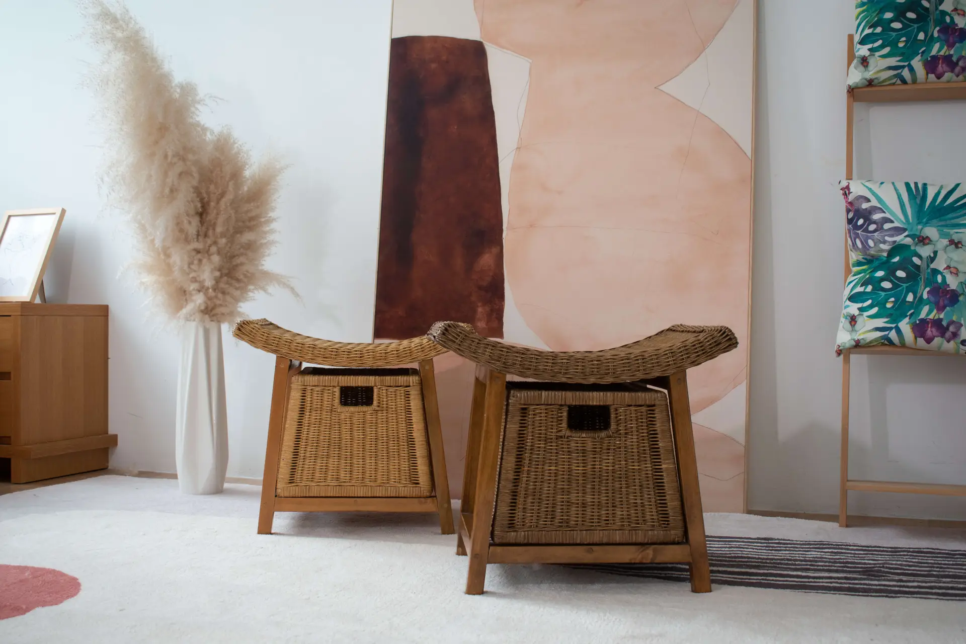 A room with two Rattan Wicker Storage Stools, a dried pampas grass arrangement in a vase, abstract wall art, and a shelf with cushions featuring colorful leaf patterns.