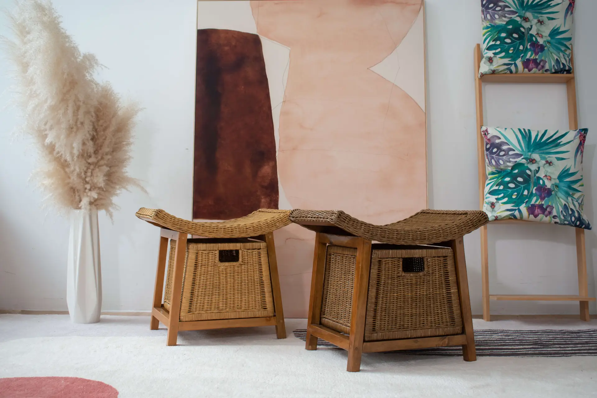 Two Rattan Wicker Storage Stools are placed in front of an abstract painting, flanked by a vase of pampas grass on one side and a ladder holding two tropical-print cushions on the other.