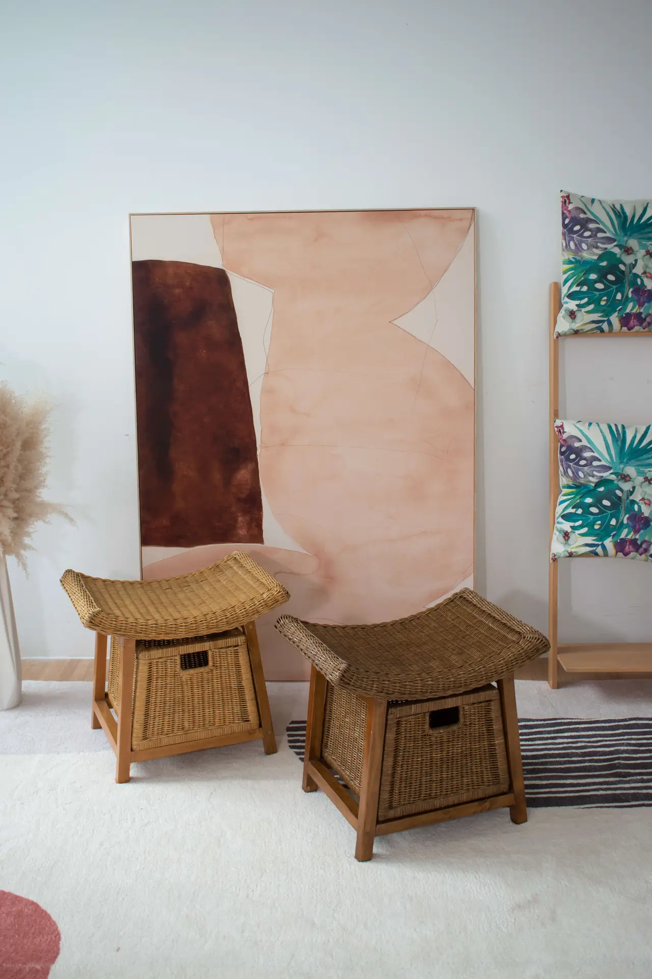 Two Rattan Wicker Storage Stools sit in front of an abstract painting leaning against a wall. Nearby, a rack displays patterned fabric, and a vase with pampas grass is placed in the corner.