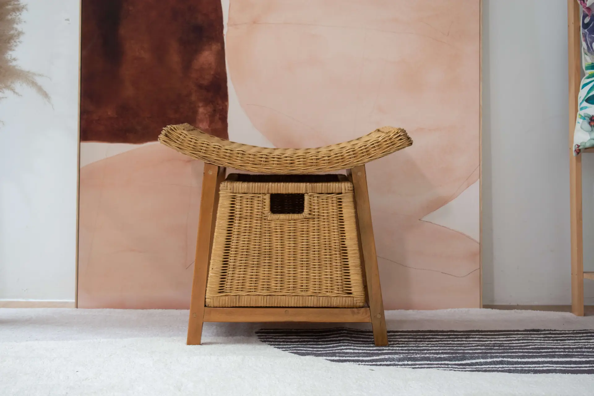 A Rattan Wicker Storage Stool with a curved seat is positioned on a rug in front of an abstract painting with shades of pink and brown.