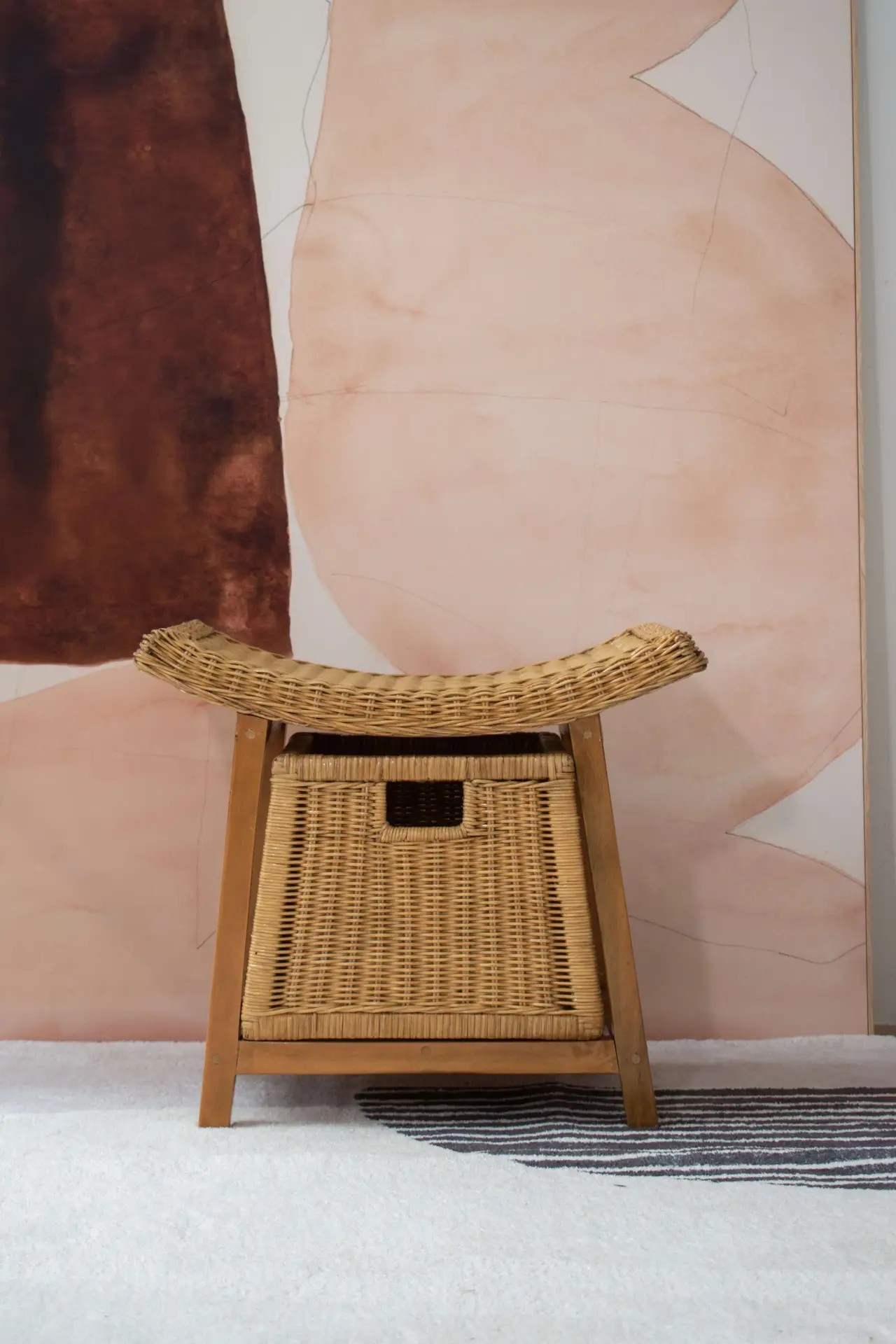 A Rattan Wicker Storage Stool and bench sit against a wall adorned with abstract pink and brown designs.