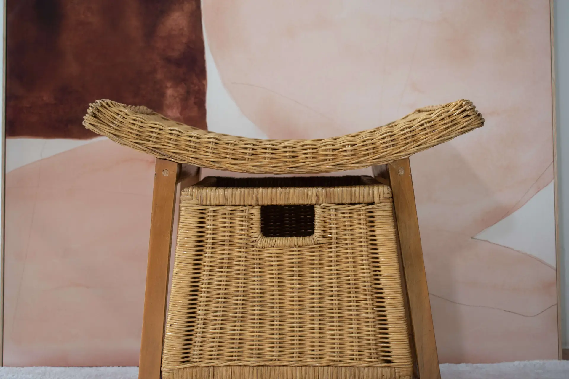A close-up of the Rattan Wicker Storage Stool with a curved backrest against a background featuring abstract brown and beige shapes.