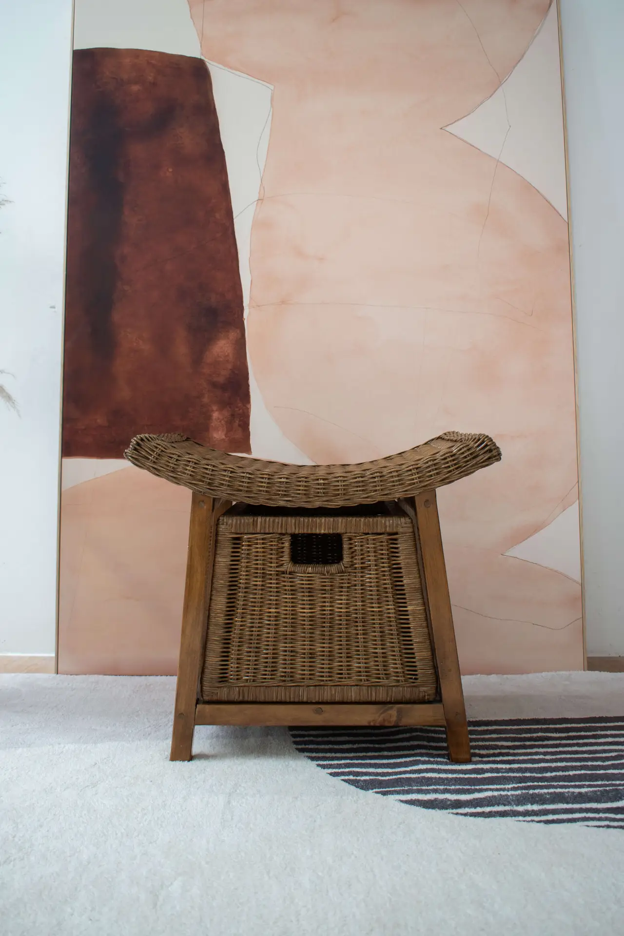 A Rattan Wicker Storage Stool with a curved seat is placed on a rug featuring striped and solid patterns, with a large abstract painting in shades of brown and peach adorning the background.