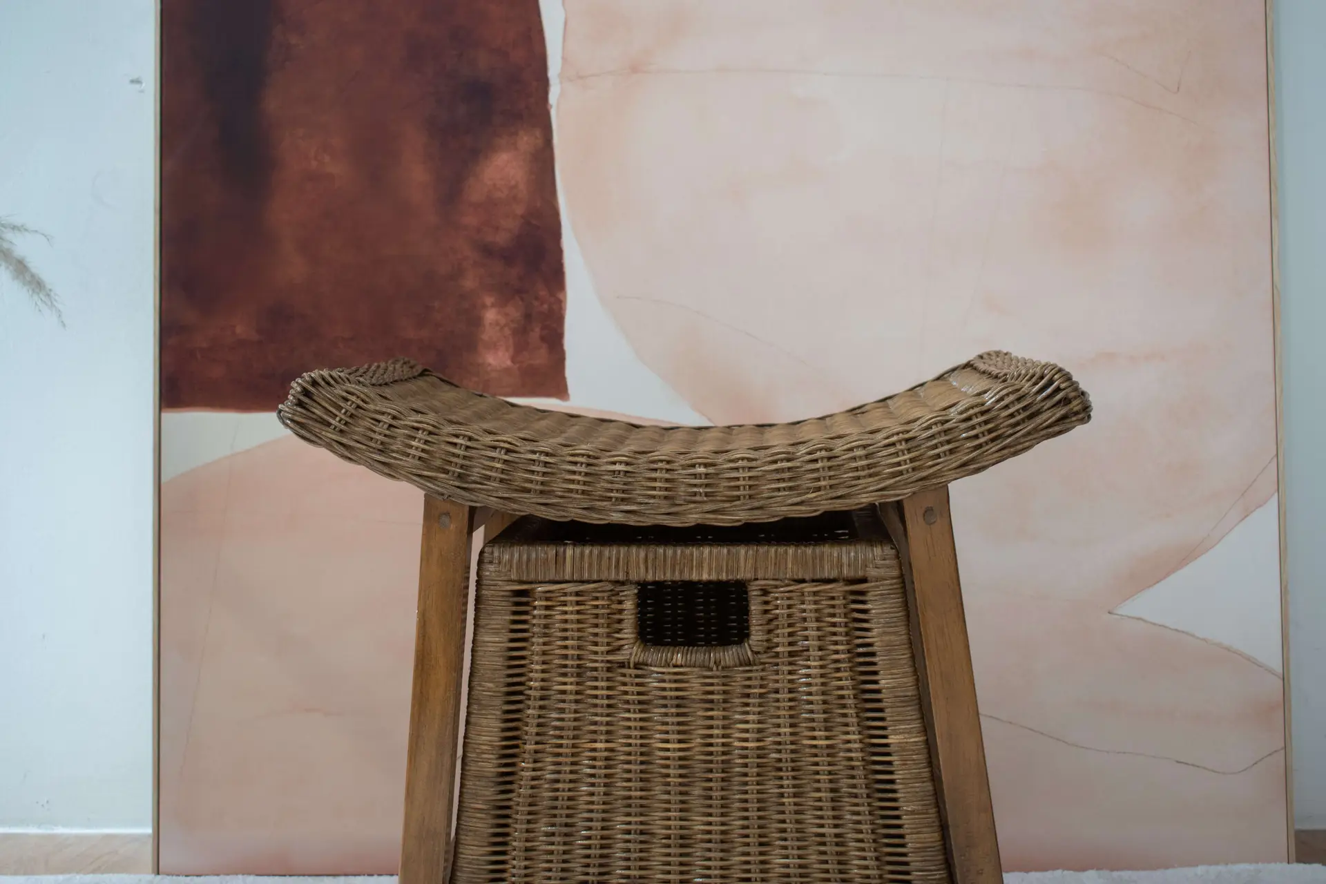 A Rattan Wicker Storage Stool sits in front of an abstract painting with brown and beige hues.