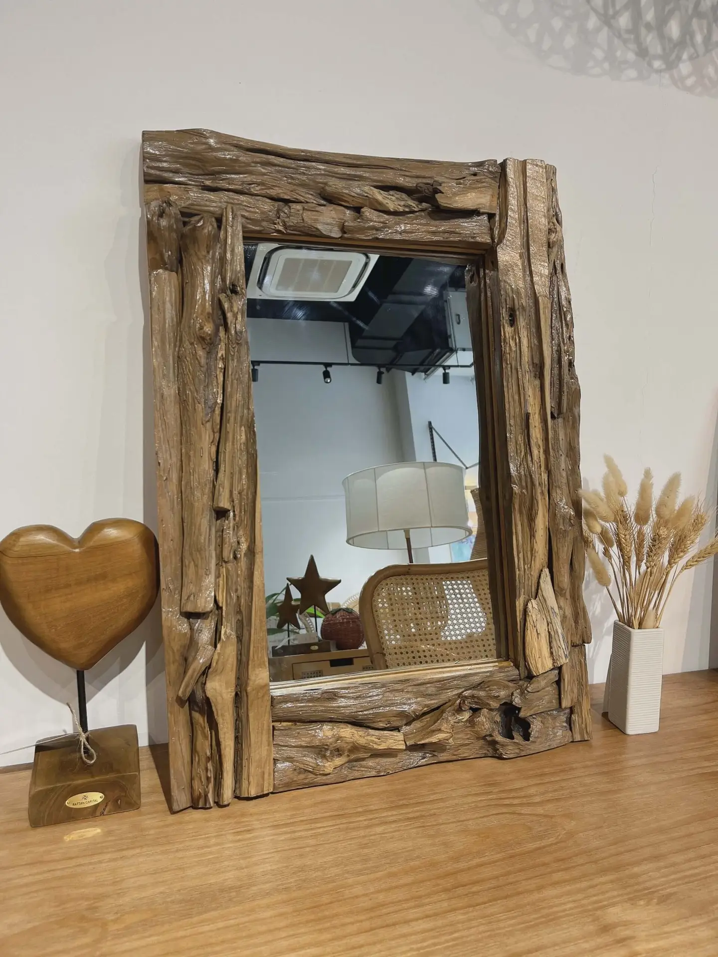 Wood-framed mirror on a wooden table with a heart-shaped lamp, dried floral arrangement in a white vase, and a woven chair visible in the reflection.