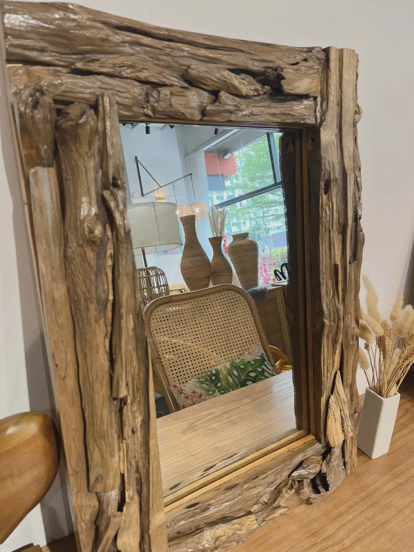 A mirror with a rustic wooden frame reflecting a room with home decor items like vases, a wicker chair, and a lamp. Dried pampas grass is arranged next to the mirror.