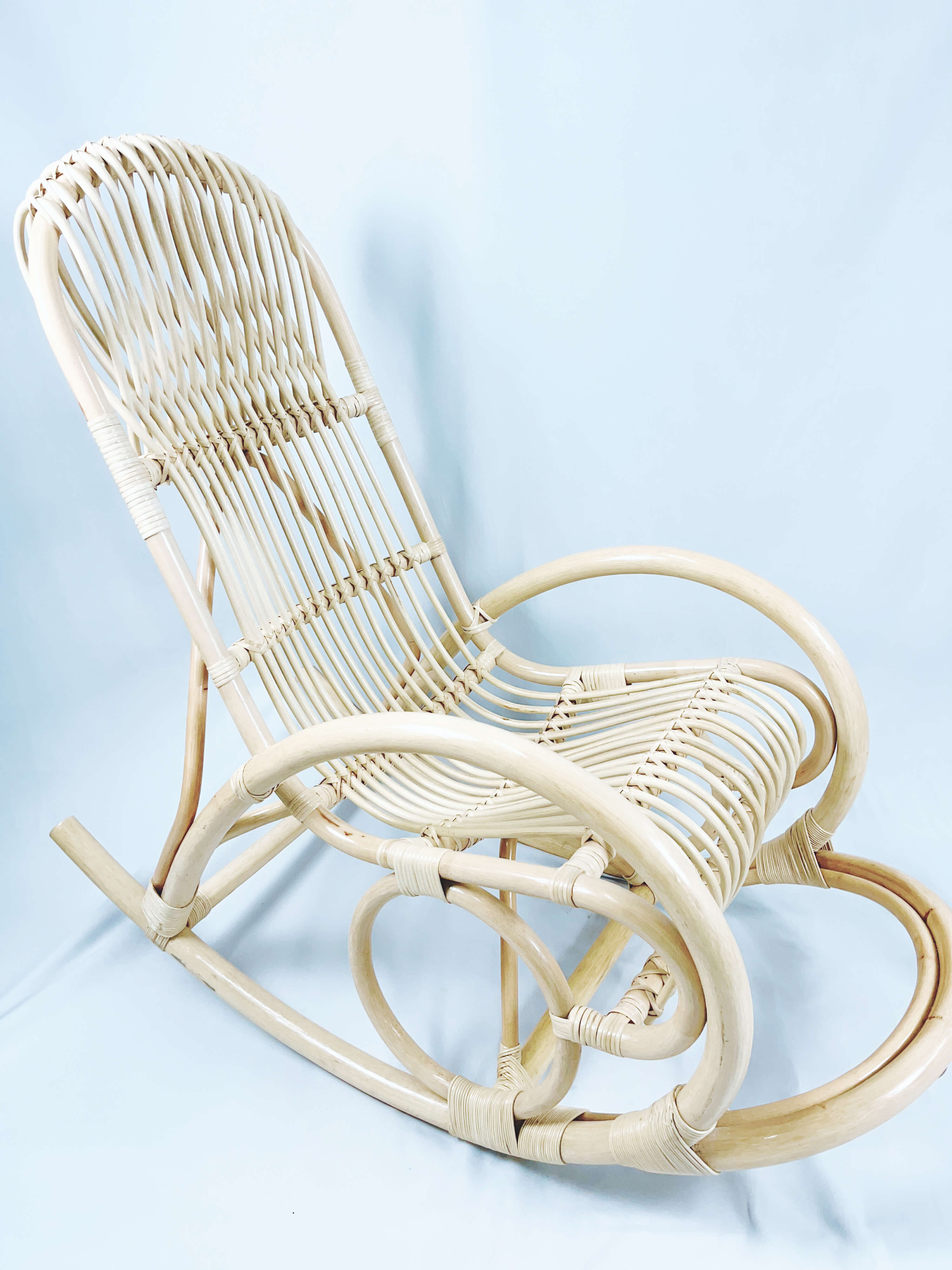 A light-colored, intricately woven wicker rocking chair with a high backrest and curved armrests, set against a plain background.