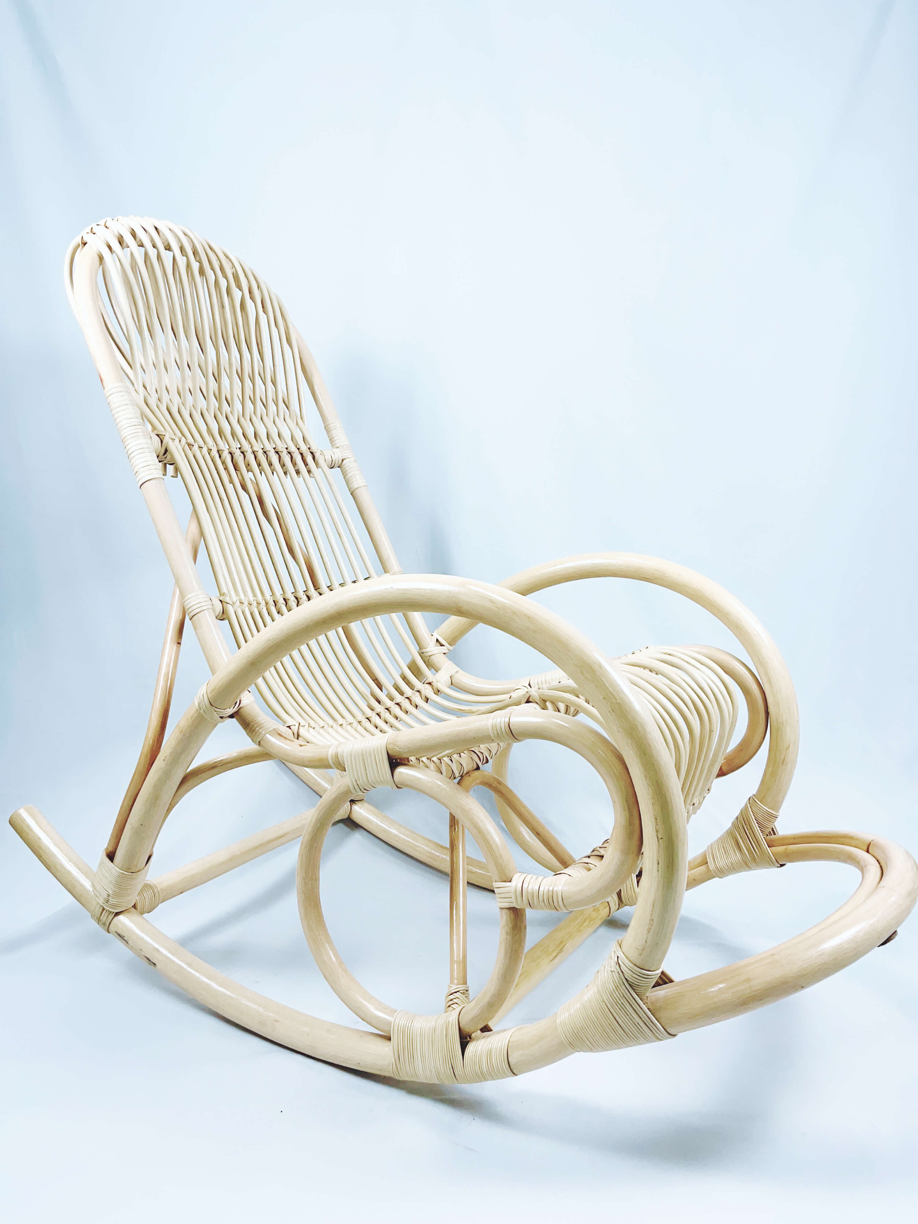 A natural-colored rattan rocking chair with curved armrests and a slatted back, set against a light background.