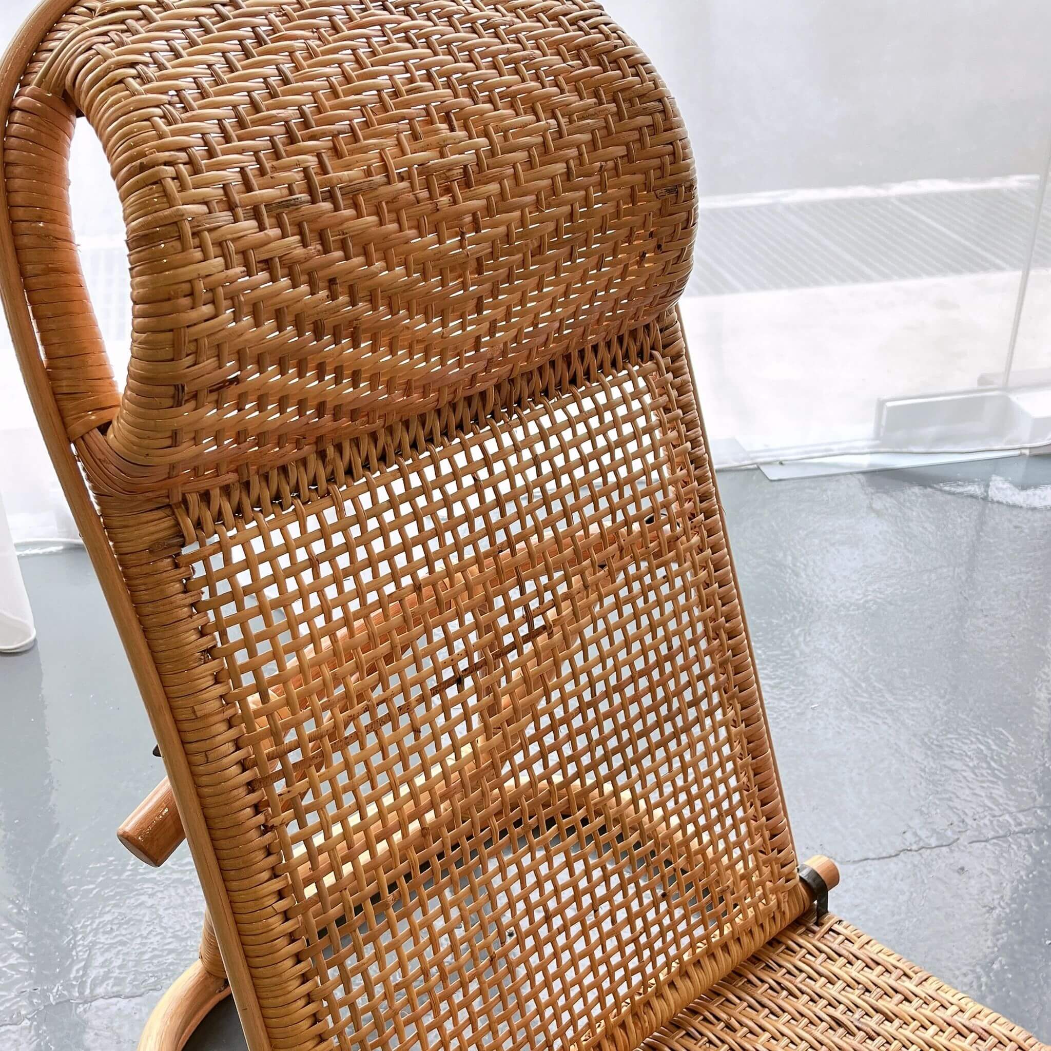 A close-up of a wicker chair with a curved backrest and a transparent acrylic background. The chair is on a shiny, gray floor.
