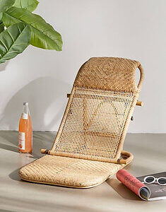 A wicker lounge chair sits on a surface with a bottle of drink, a plant, a pair of sunglasses, and an open magazine nearby.