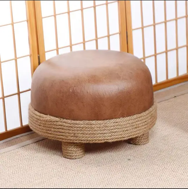 A round brown ottoman with a leather-like texture and rope detail at the base, placed on a beige carpet beside wooden sliding doors.