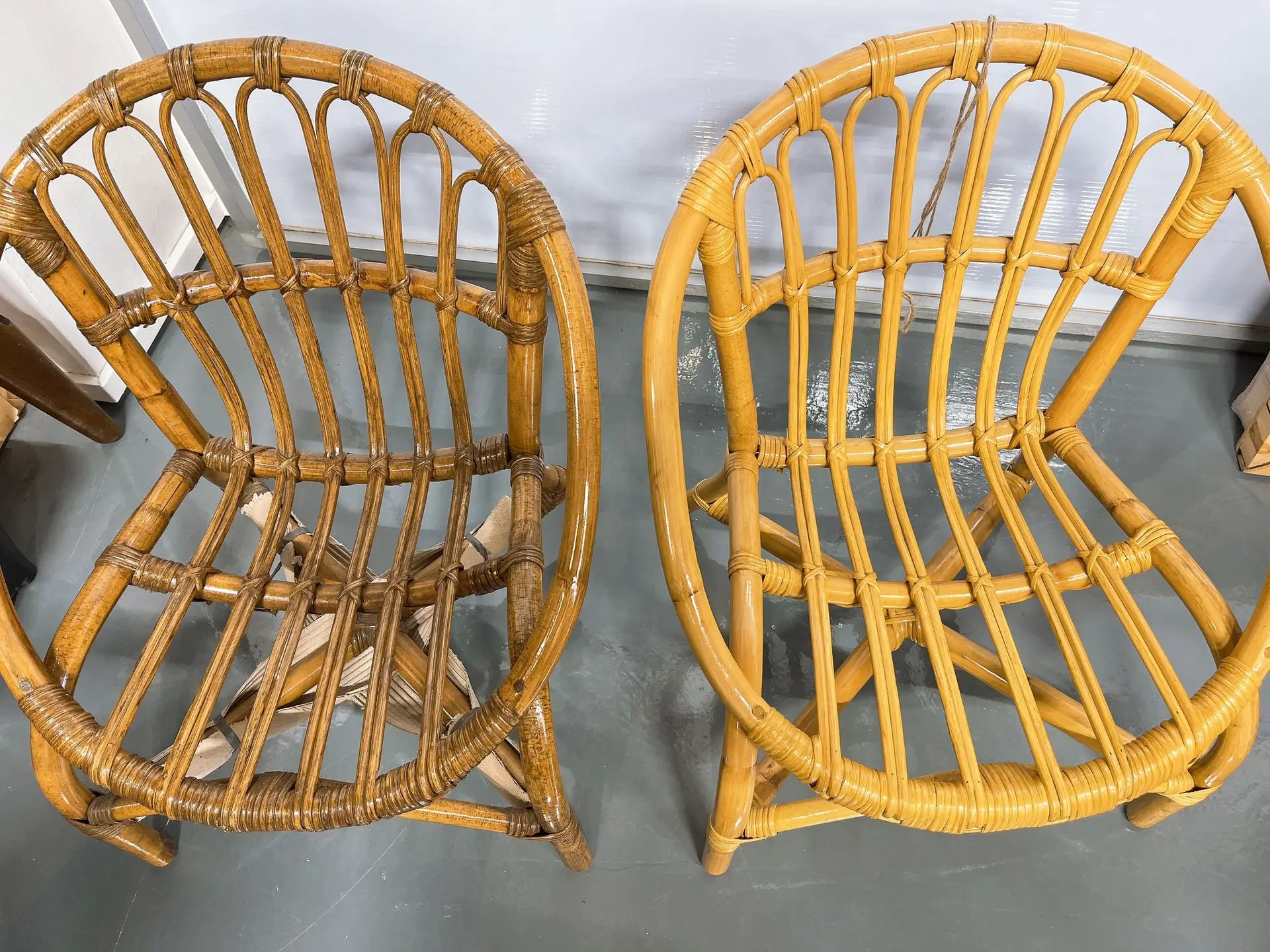 Two rattan wicker chairs with curved backs and seats are positioned side by side on a gray floor in front of a white wall. The chair on the left has a piece of wood underneath the seat.