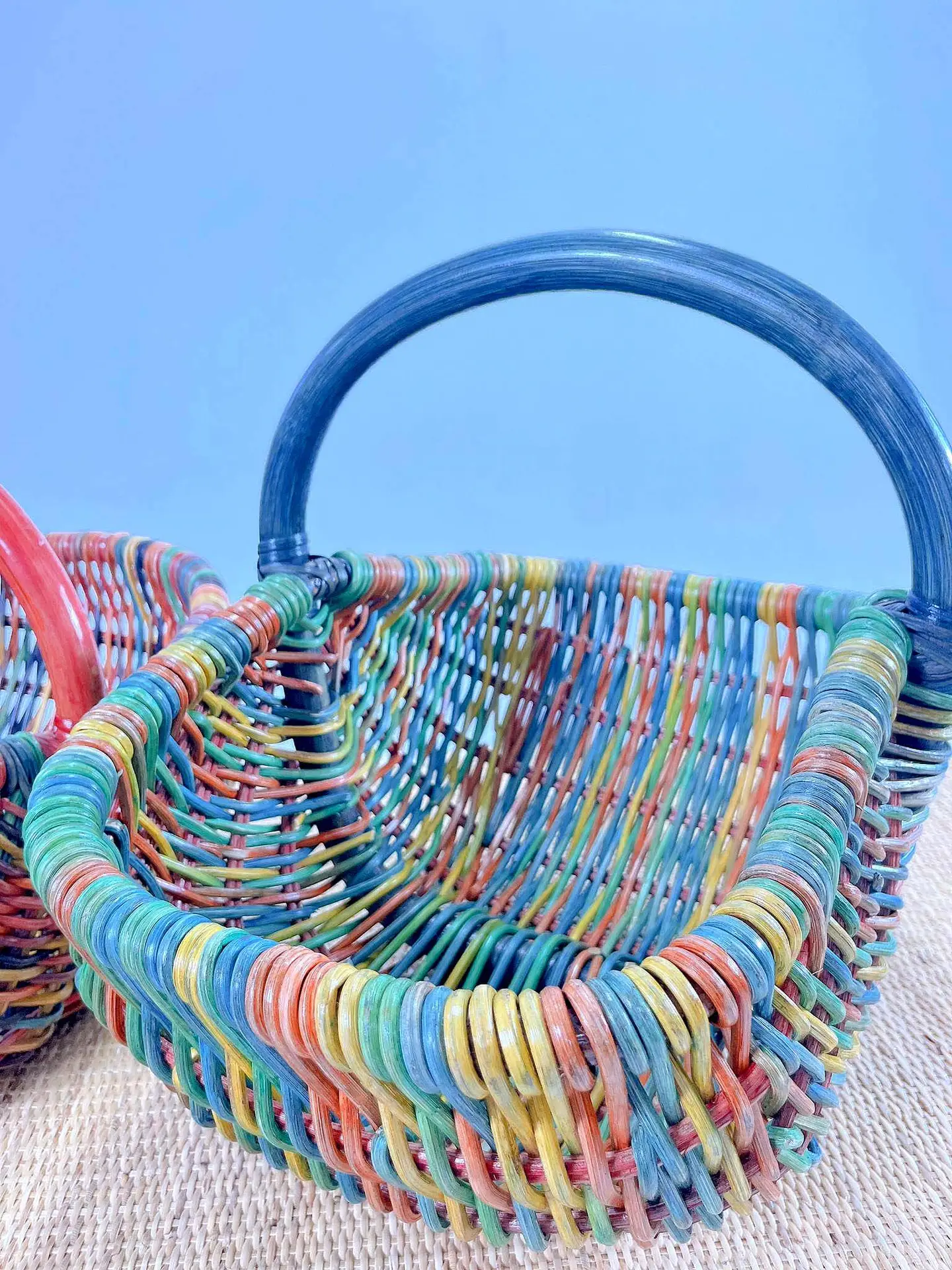 Close-up of two Rainbow Aristide Baskets with handles, showcasing an array of blue, green, red, and yellow hues, set on a textured surface against a light blue background.