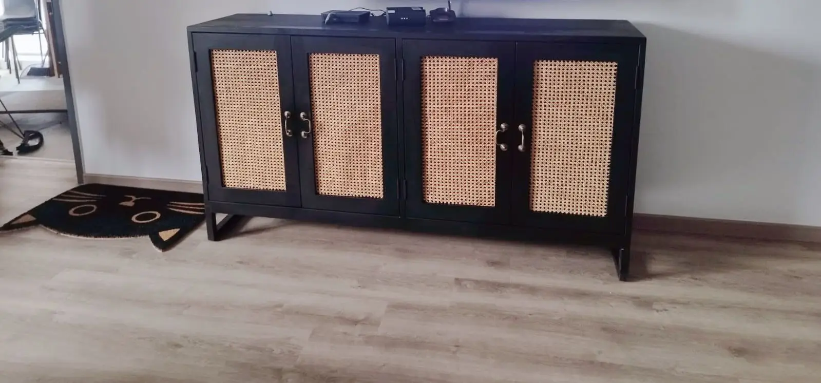 A Pacific Rattan Cabinet with four woven rattan panel doors stands against a wall. On top of it are two small electronic devices. A cat-shaped mat is on the floor to the left of the cabinet.