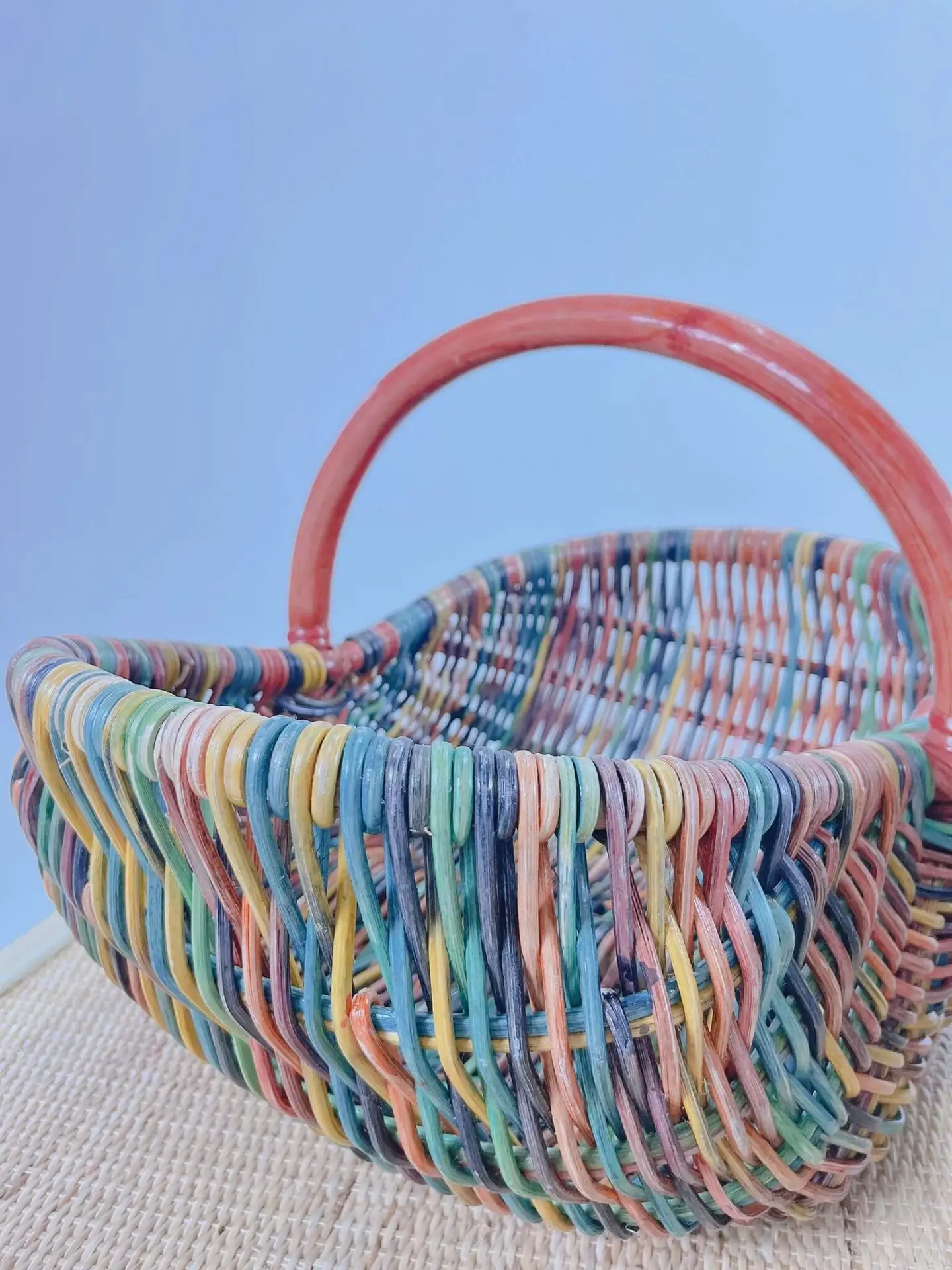Dawn's Plant Pot, characterized by its multicolored woven design and a reddish handle, sits on a beige mat against a light blue background.