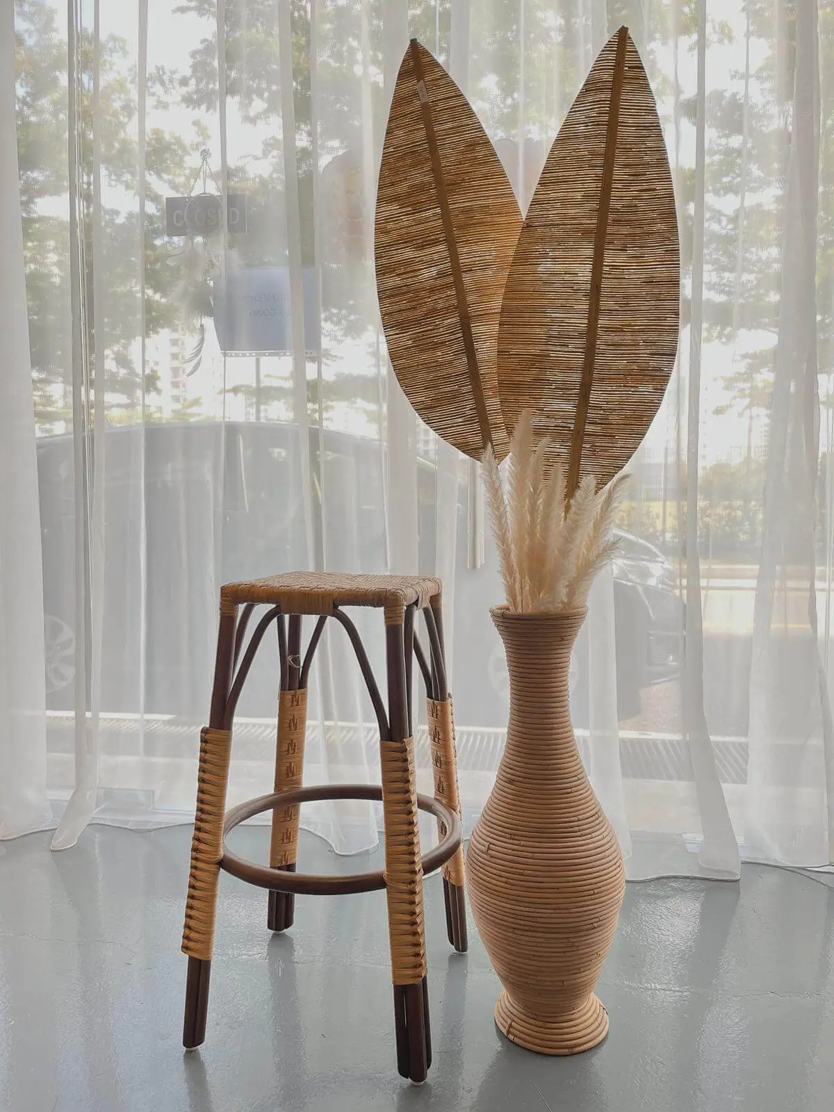 A wicker stool is placed beside a tall, woven vase containing dried pampas grass and two large, leaf-shaped wicker decorations. The scene is set in front of a sheer white curtain with a view outside.
